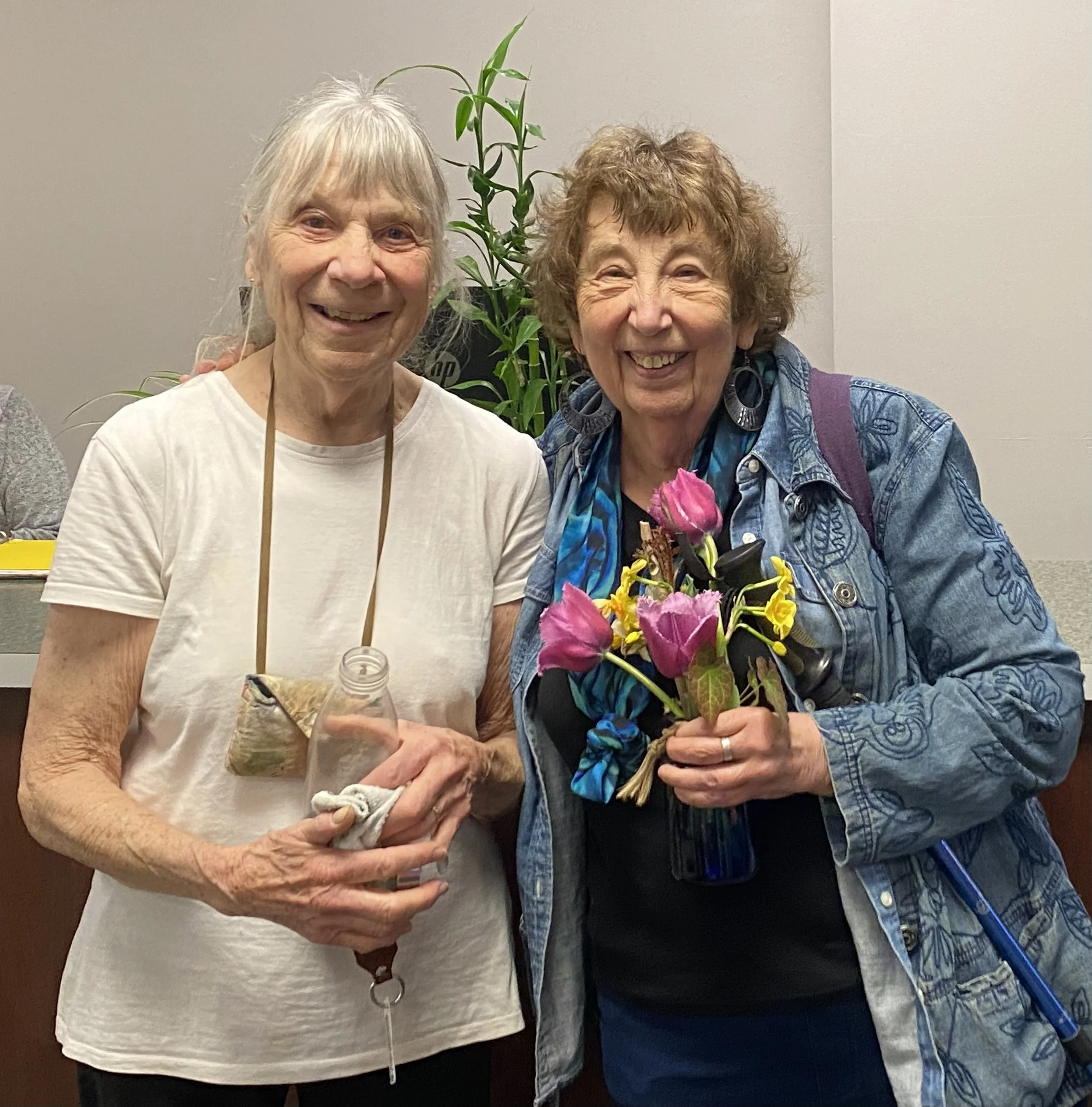 Two members smiling, one holding a bouquet of pink and yellow flowers, standing in front of a plain wall.