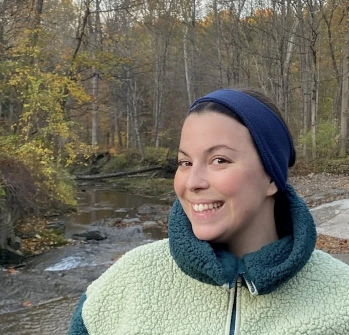 Picture of Leah standing in front of a stream in the fall, wearing a fluffy sweatshirt.