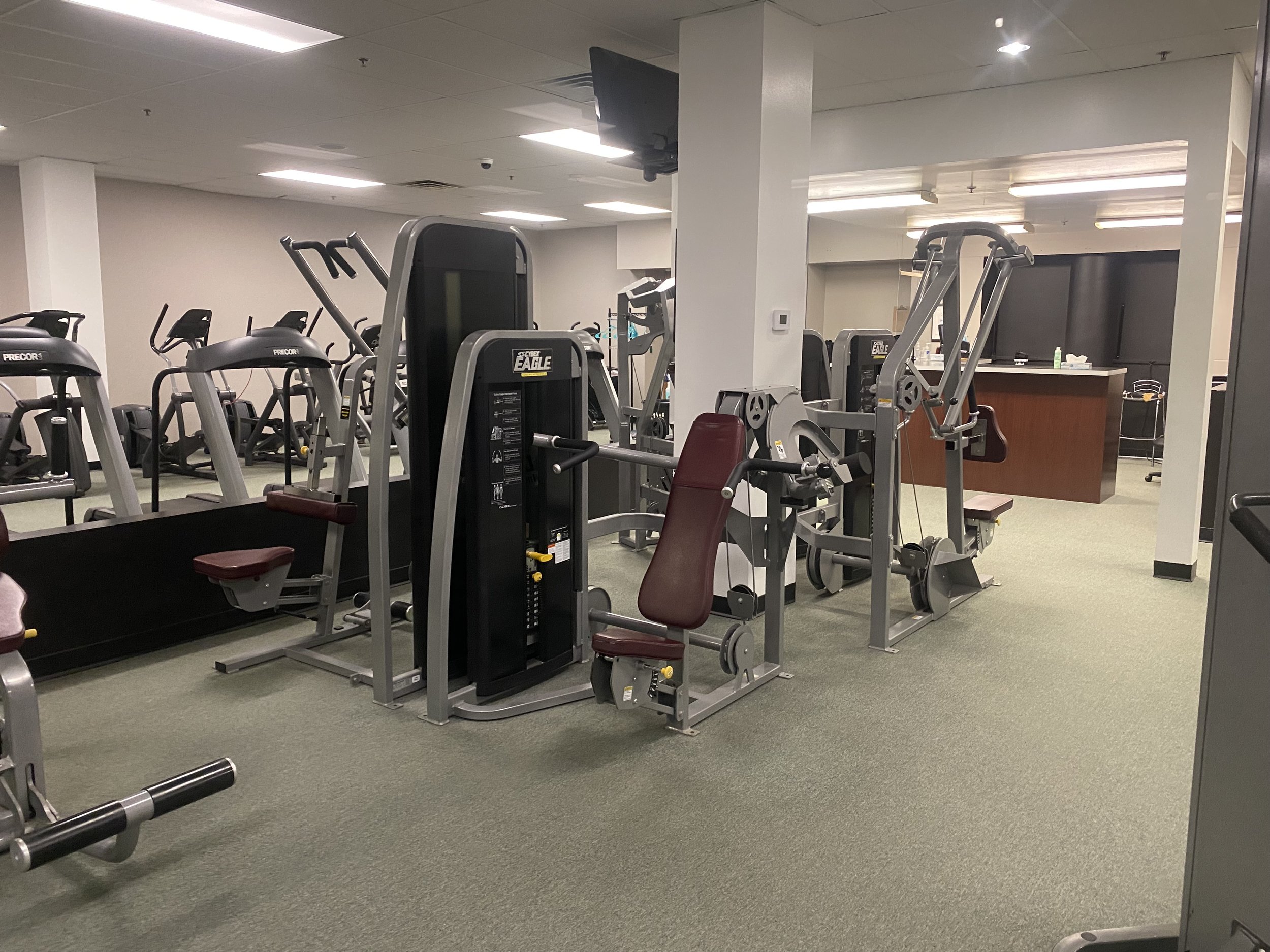 Fitness gym with various workout equipment, including weight machines and cardio machines, in an indoor space with bright lighting and neutral-colored walls and floors.