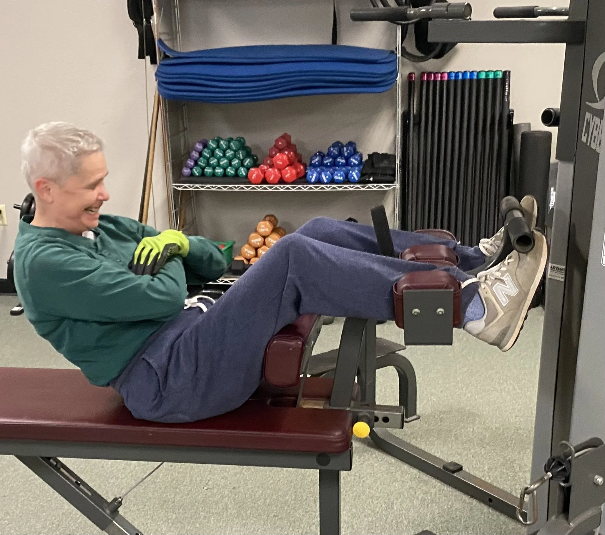 Person sitting on a workout bench in a gym, smiling and wearing green and black workout clothes, with a shelf of colorful kettlebells and resistance bands in the background.