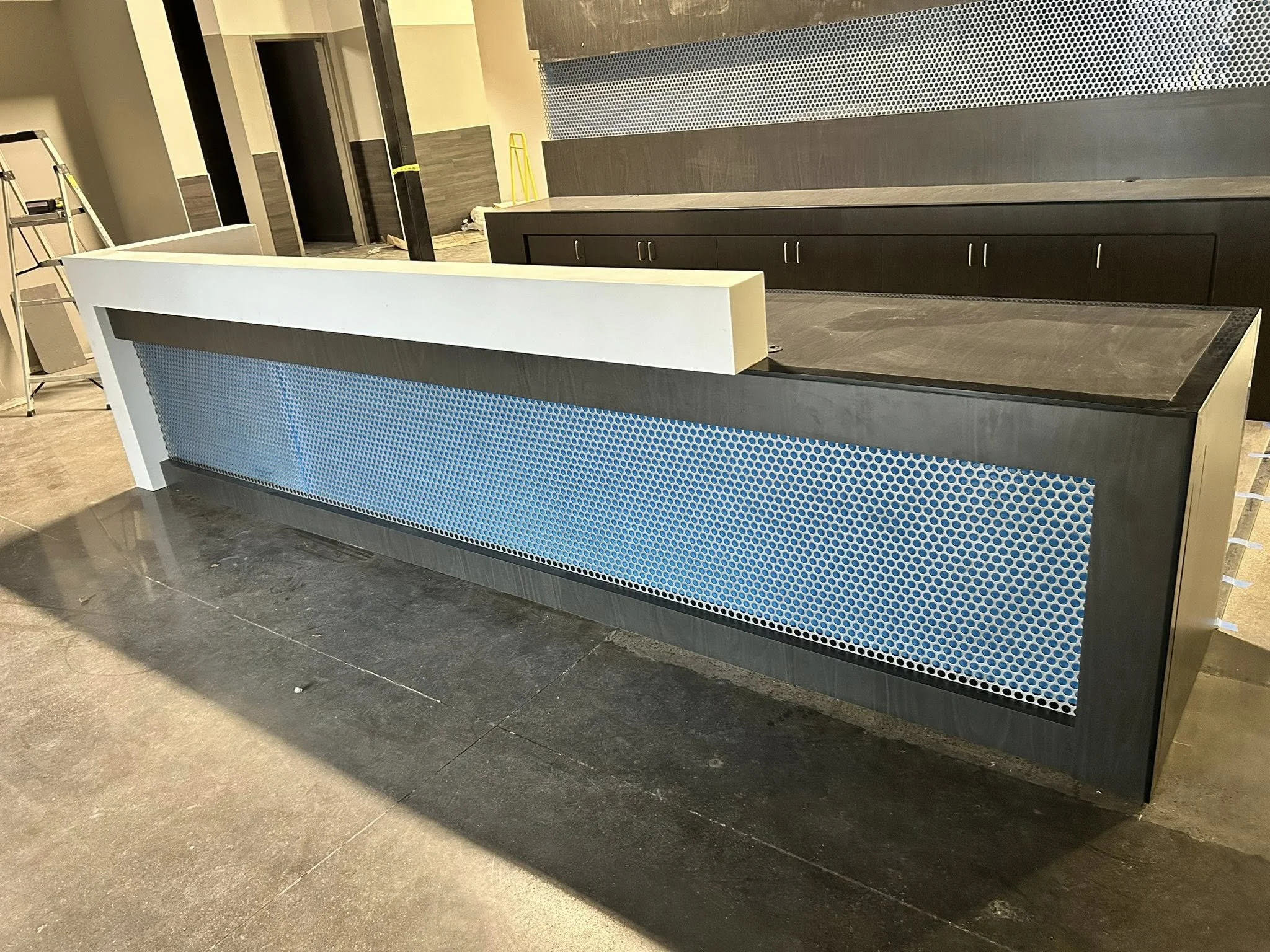 Construction area with a black counter with blue perforated metal front, dark wood cabinets, and construction tools such as a ladder and a yellow level in the background.