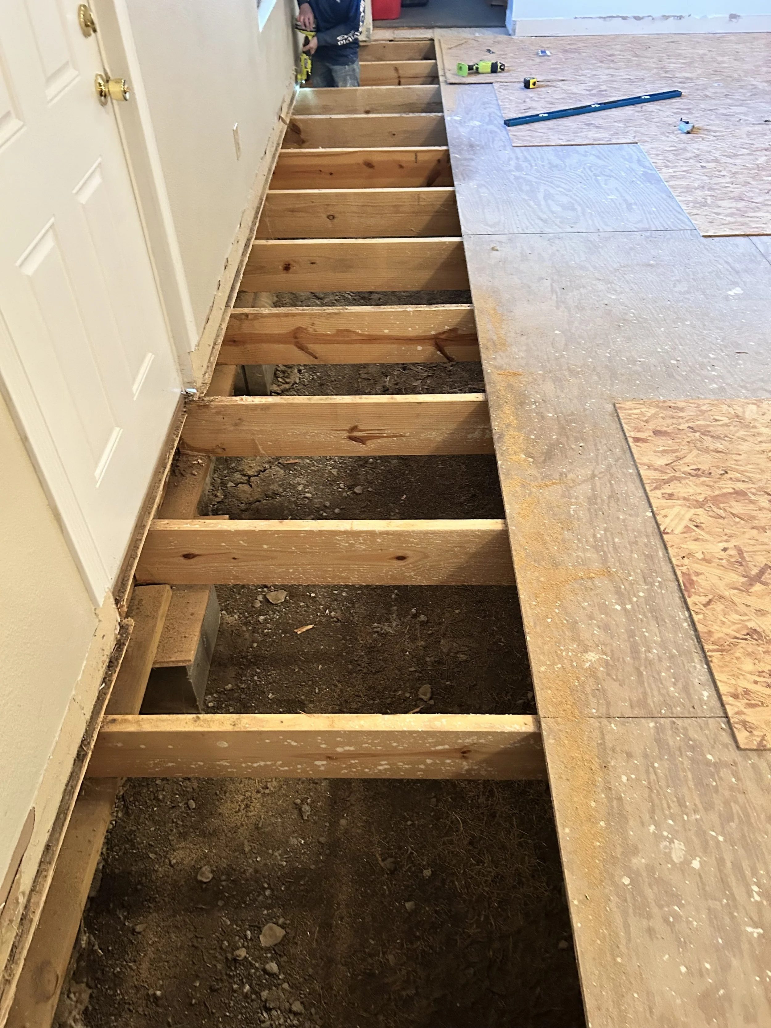 Interior of a house under renovation, showing a floor with removed flooring and exposed wooden subfloor and joists. A person is working with a drill, and tools are scattered on the plywood sheet.