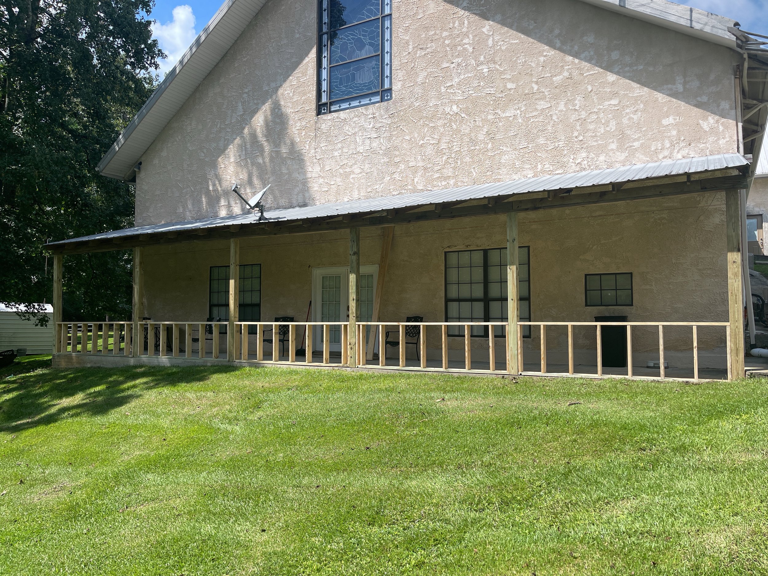 The image shows the back of a two-story house with beige textured walls and a gabled roof. There is a balcony with a railing, underneath which are four windows and a door. The house has a porch with wooden supports, a roof, and a satellite dish attached to it. The yard has green grass and is partly shaded by trees.