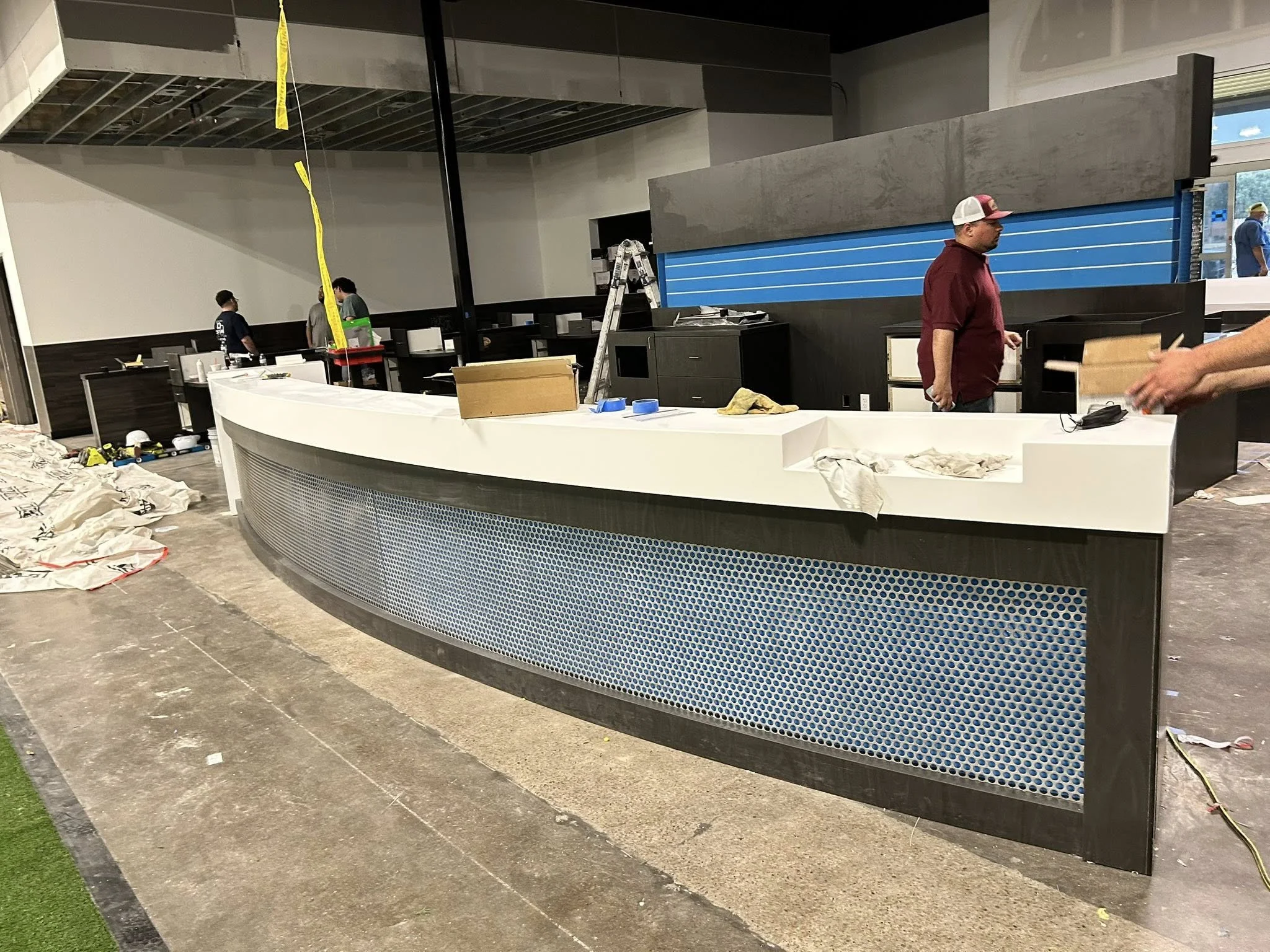Interior of a building under construction or renovation. A curved reception desk in the foreground with construction tools and materials on it. A ladder and workers are visible in the background. Walls and ceiling are unfinished.