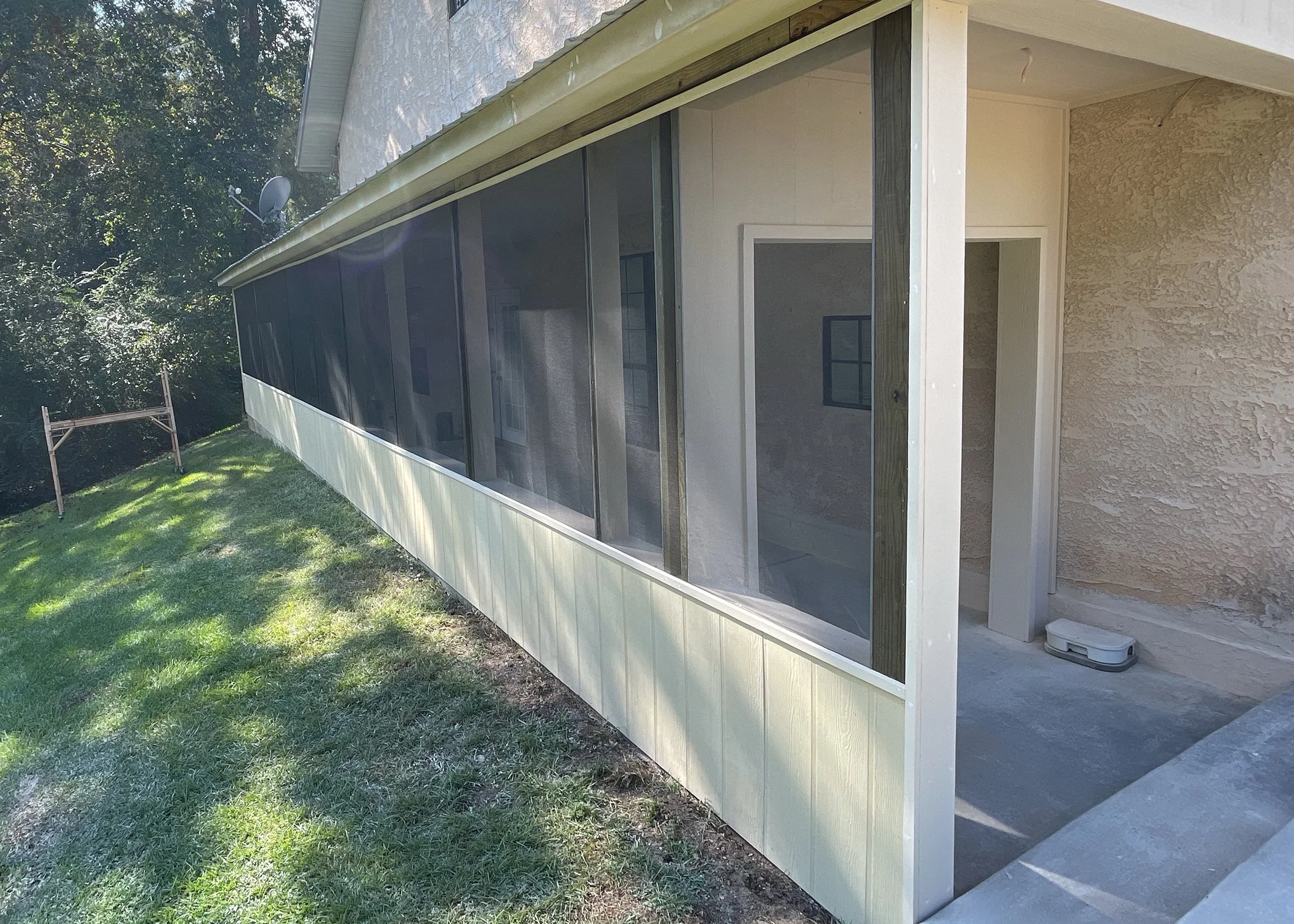 Side view of a house with a screened porch, light-colored exterior walls, and a small step leading to the porch area. There are green trees and grass surrounding the house.