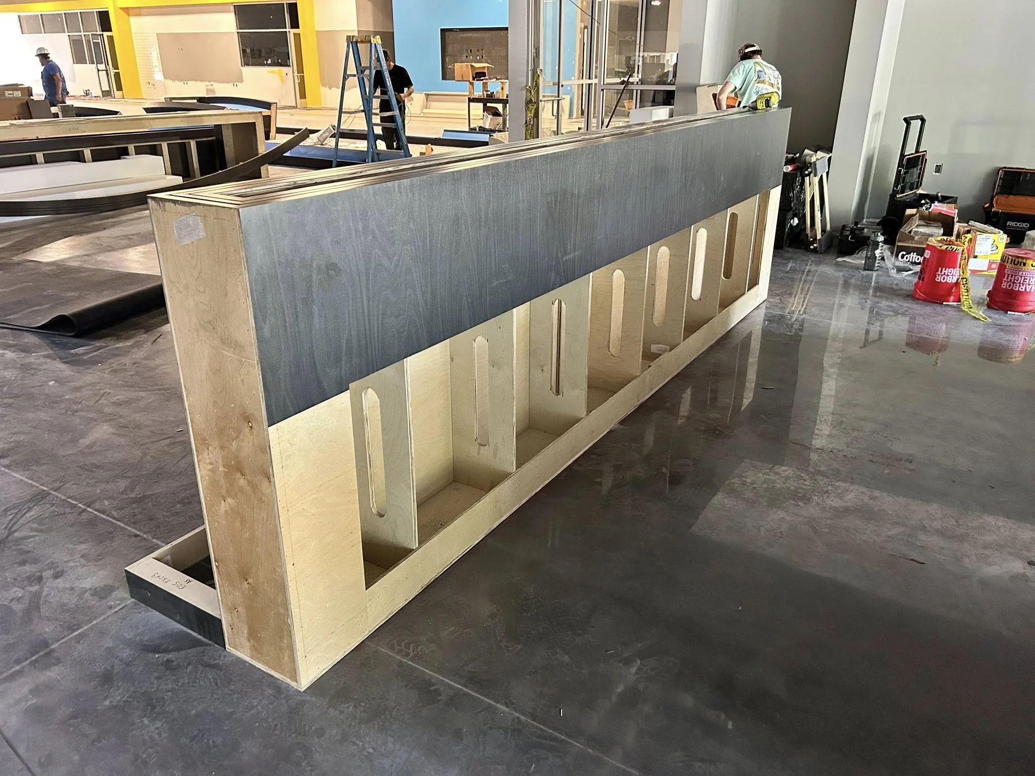 Construction workers assembling a wooden divider or booth in an indoor space, with construction tools and supplies around.