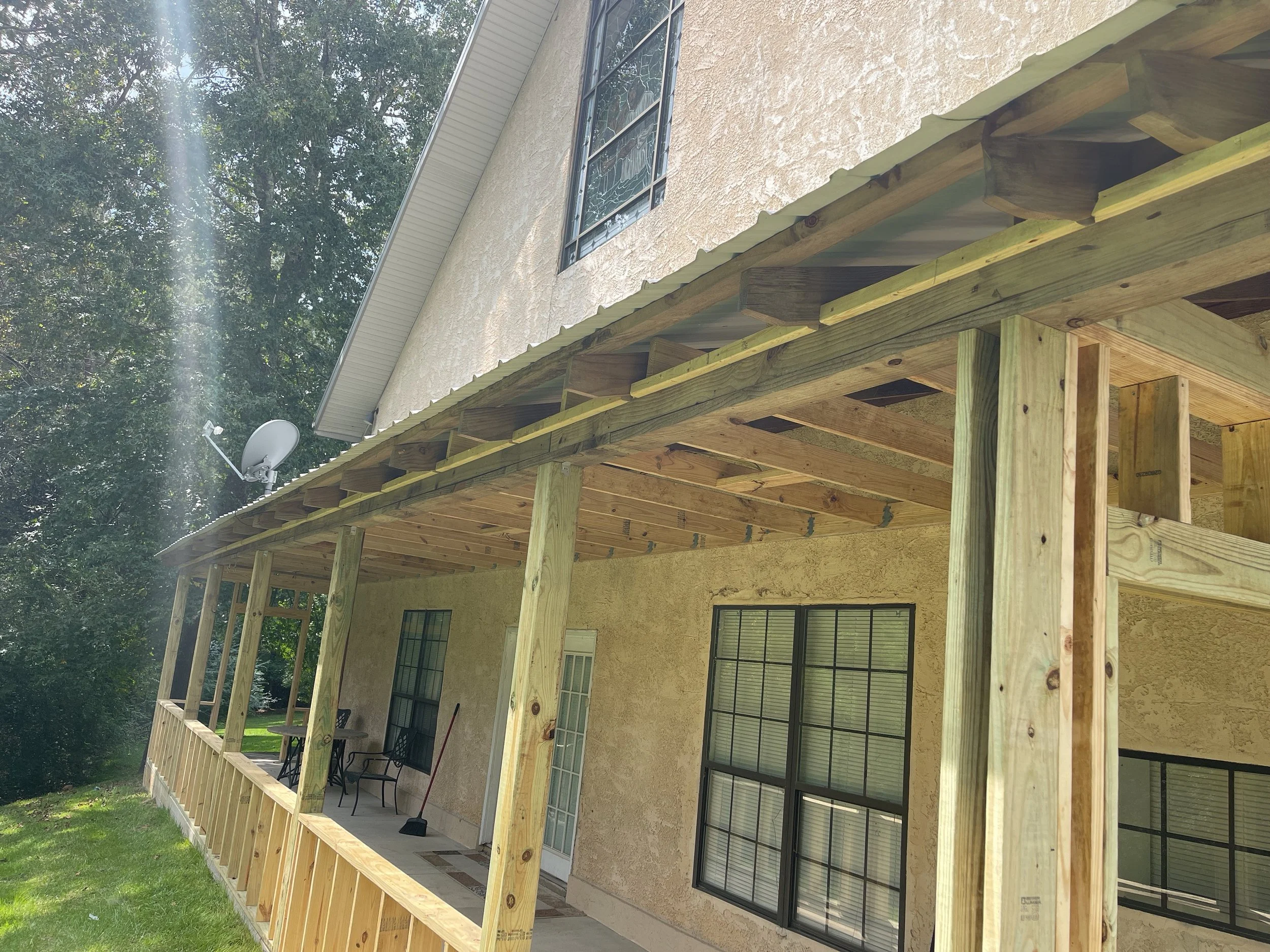 A house under construction with a new wooden balcony and railing, sitting on a grassy lawn with trees in the background, satellite dish attached to the house, and windows with black frames.