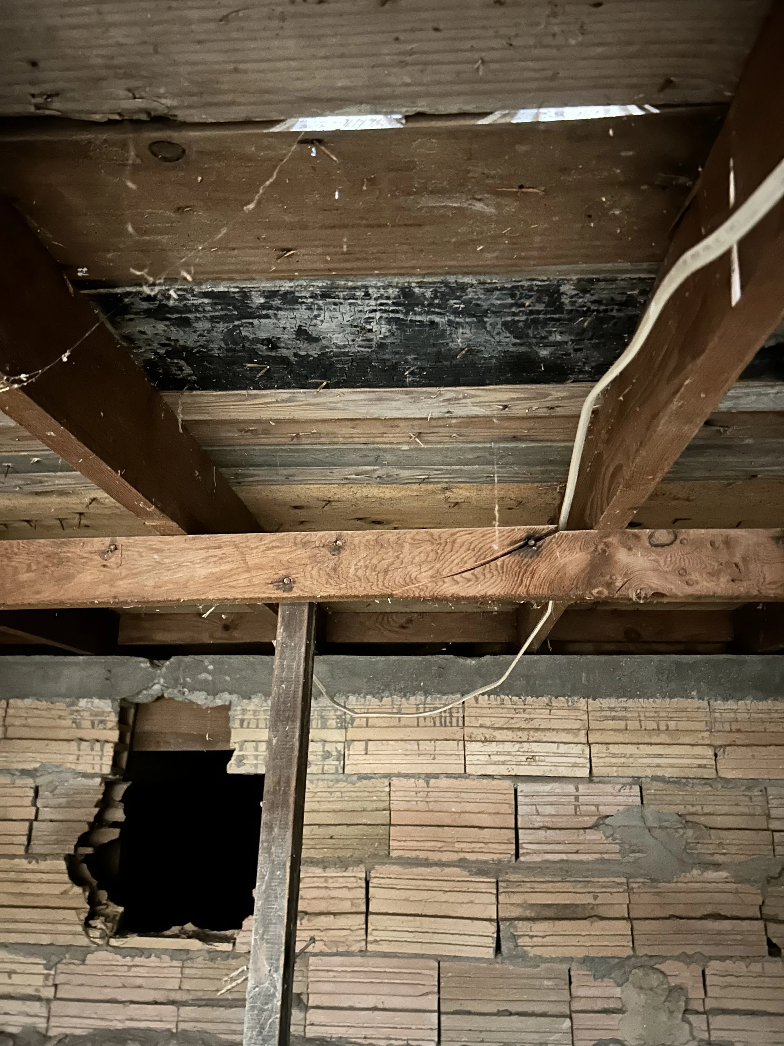 Construction area with wooden beams, brick wall, and visible hole in the ceiling, possibly for plumbing or electrical work.
