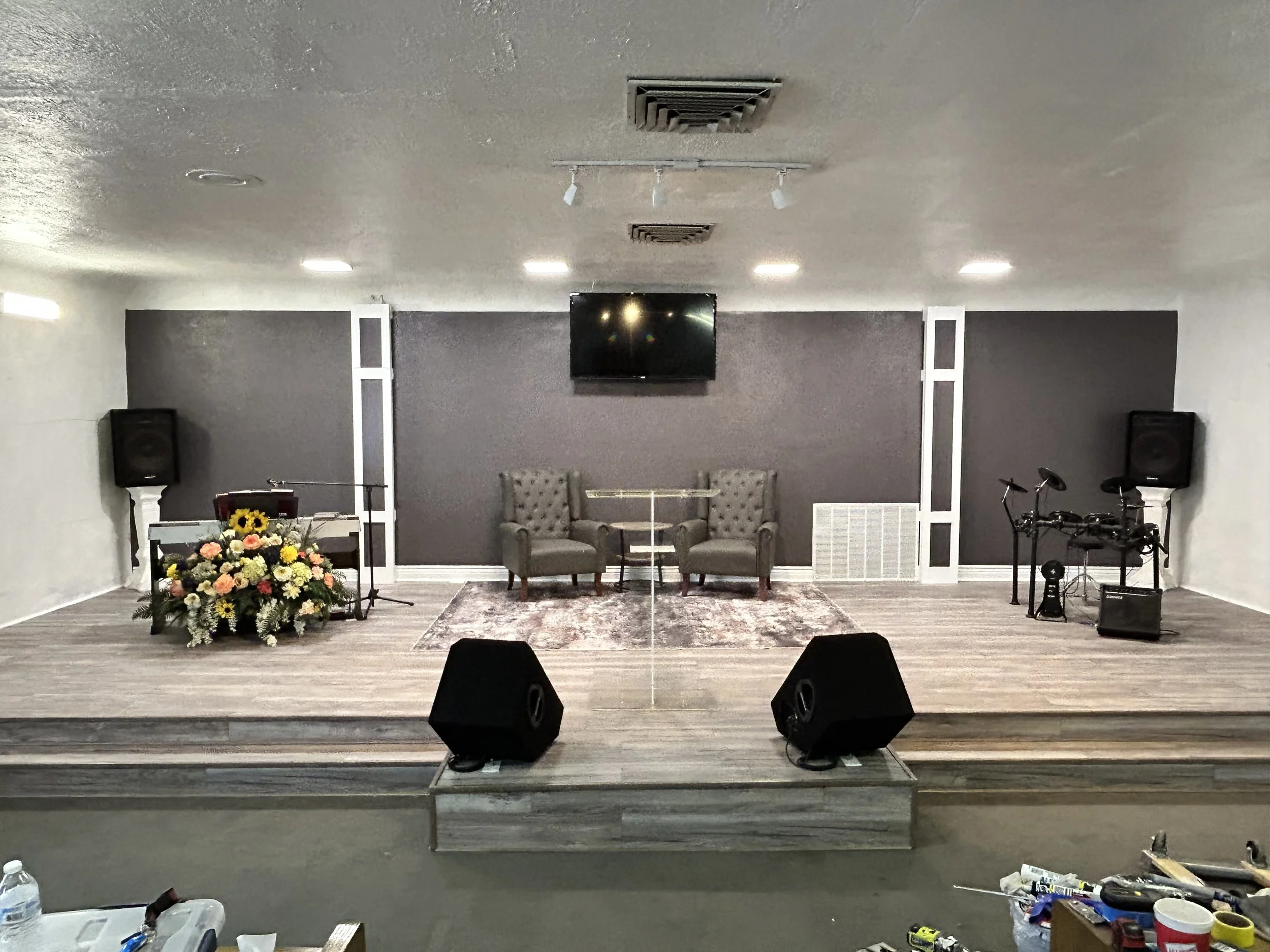 Stage with two chairs, a bouquet of flowers on a stand, and musical equipment, including speakers, microphones, and electronic drums.