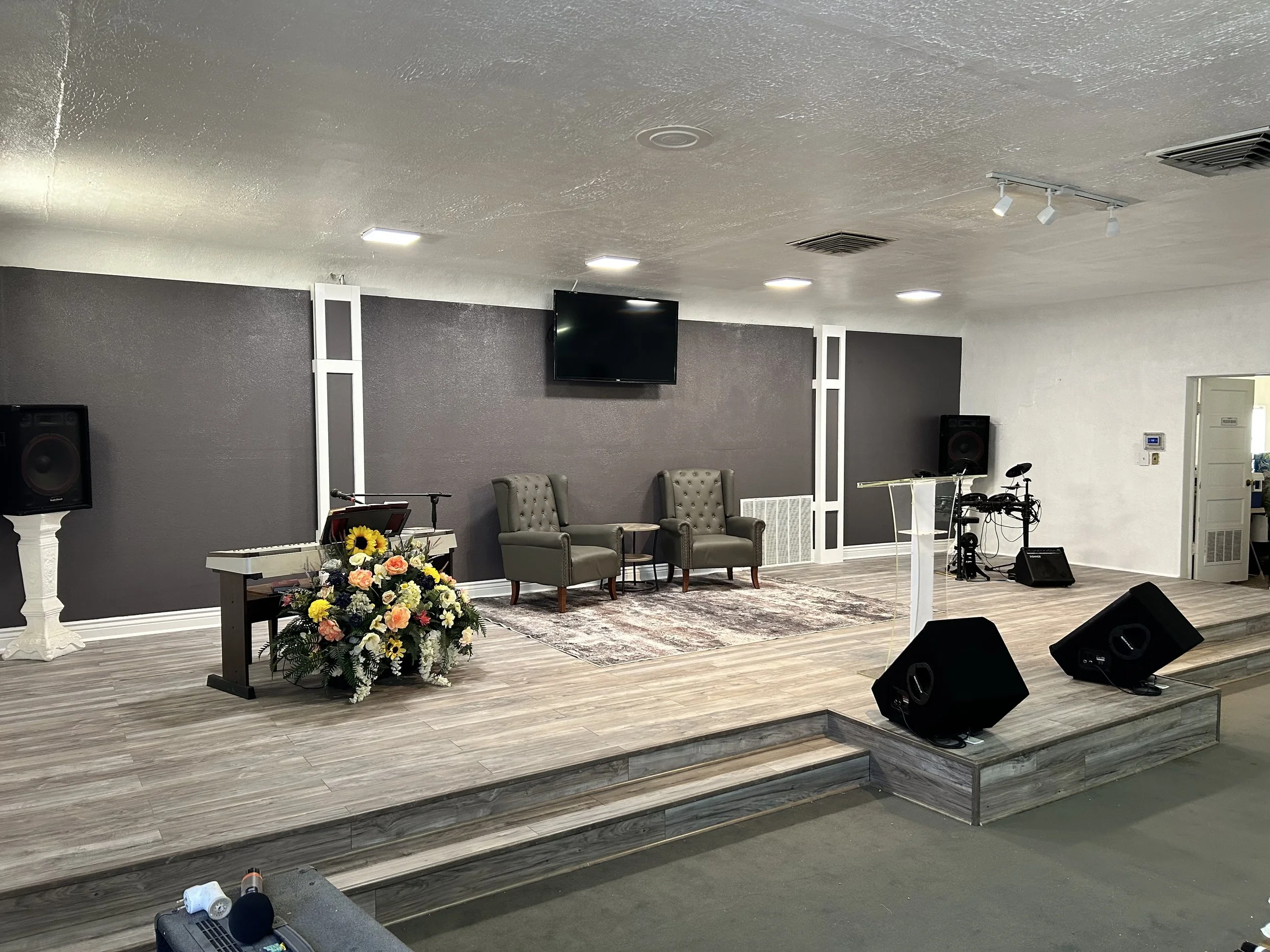 A stage with a flower arrangement, chairs, musical instruments, and speakers in a room with a textured gray wall and ceiling lights.