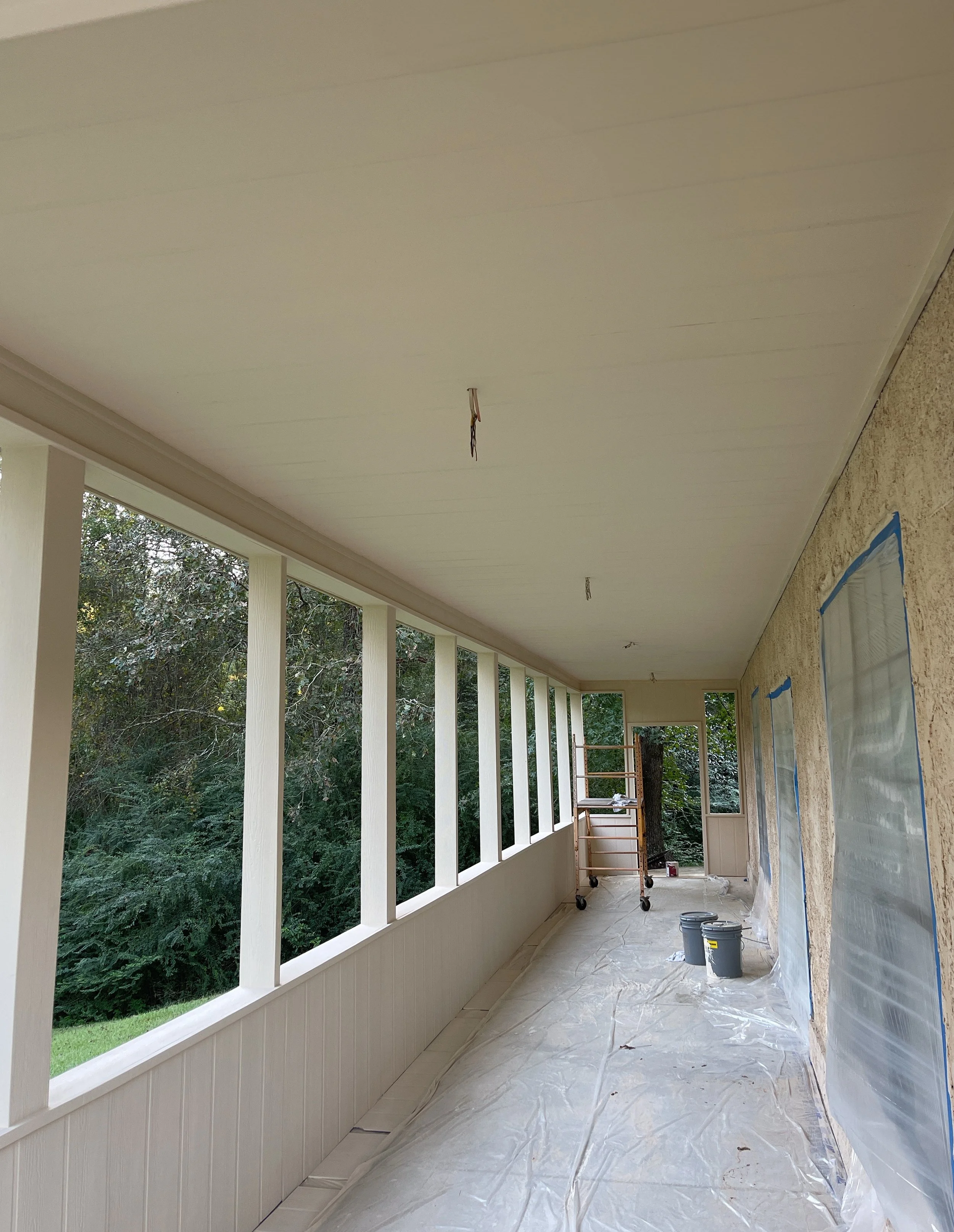 Porch under construction with white paneling on ceiling, white railing, windows covered with plastic, and painting supplies on the floor.