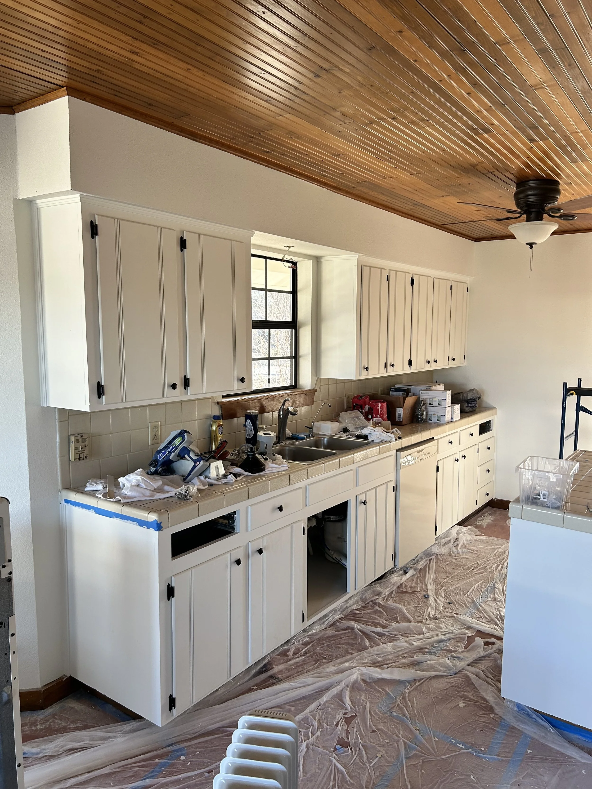 Kitchen undergoing renovation with cabinets removed, tools, and protective plastic on the floor.