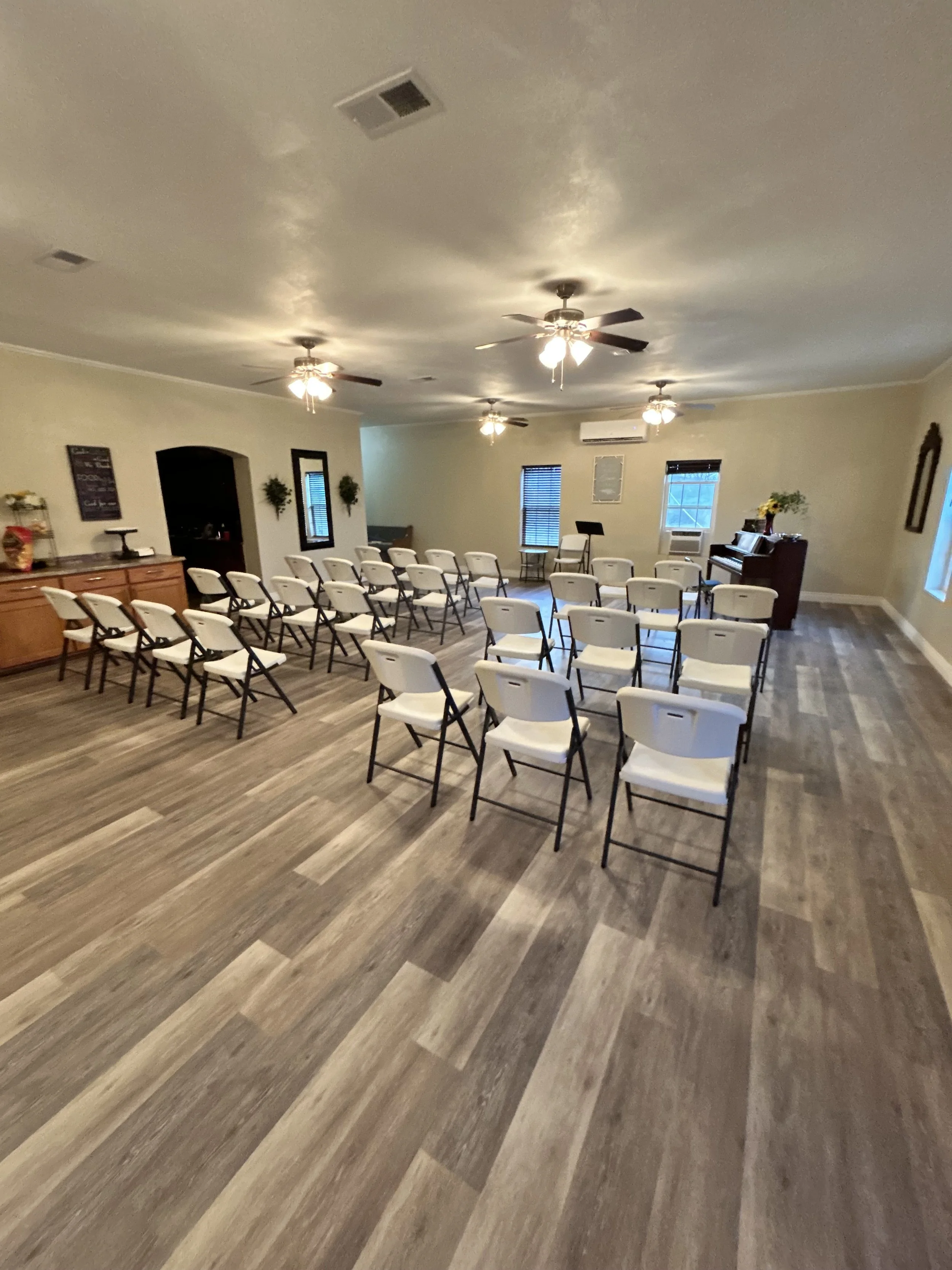 An indoor room setup with rows of white folding chairs facing a small stage area. The room has a wooden floor, ceiling fans, and windows with blinds. There is a piano against the right wall, and some decorations including plants and framed pictures.