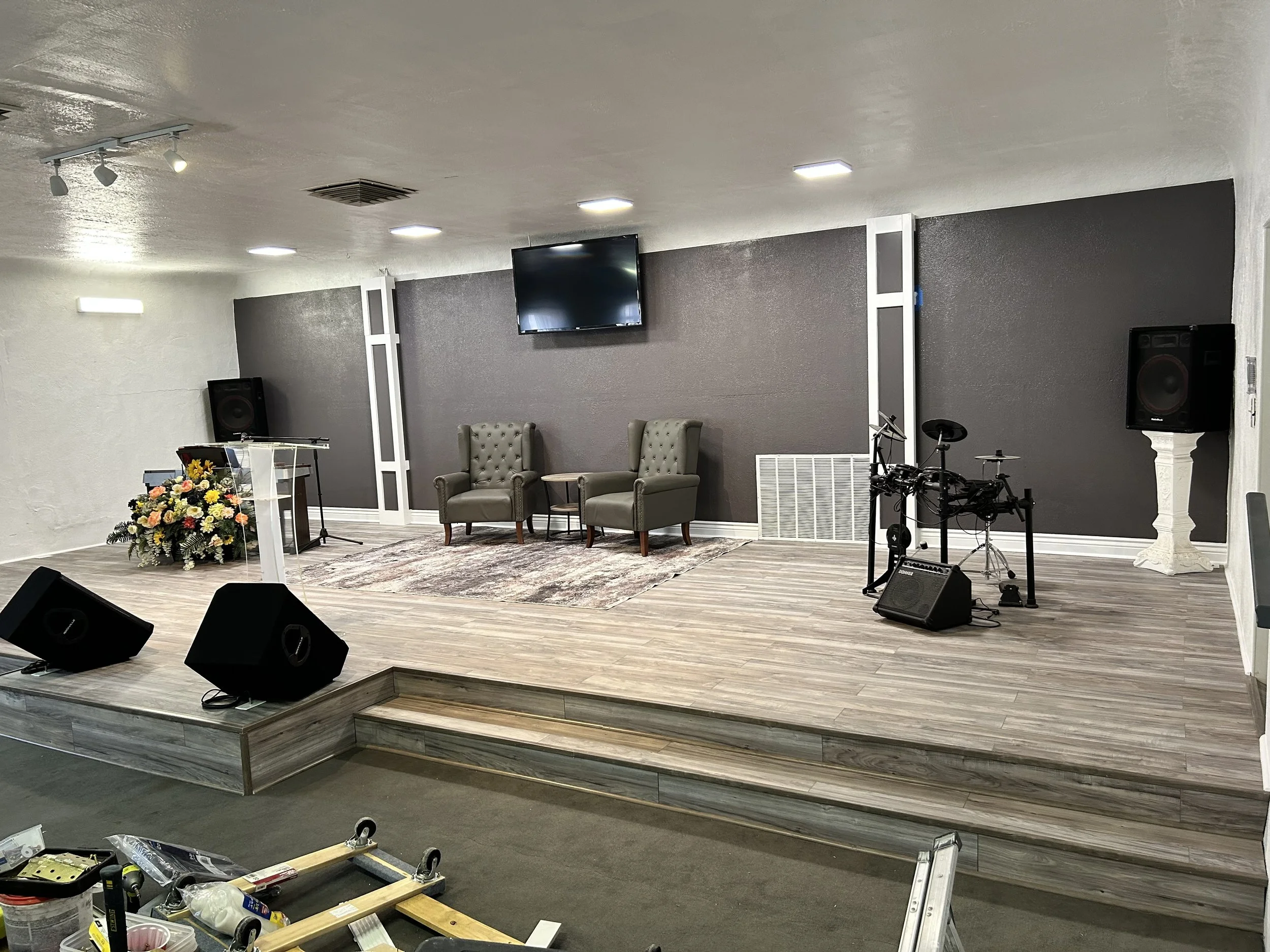 Stage with a flower arrangement, two beige chairs, a large black screen on the wall, musical equipment, and construction tools in the foreground.