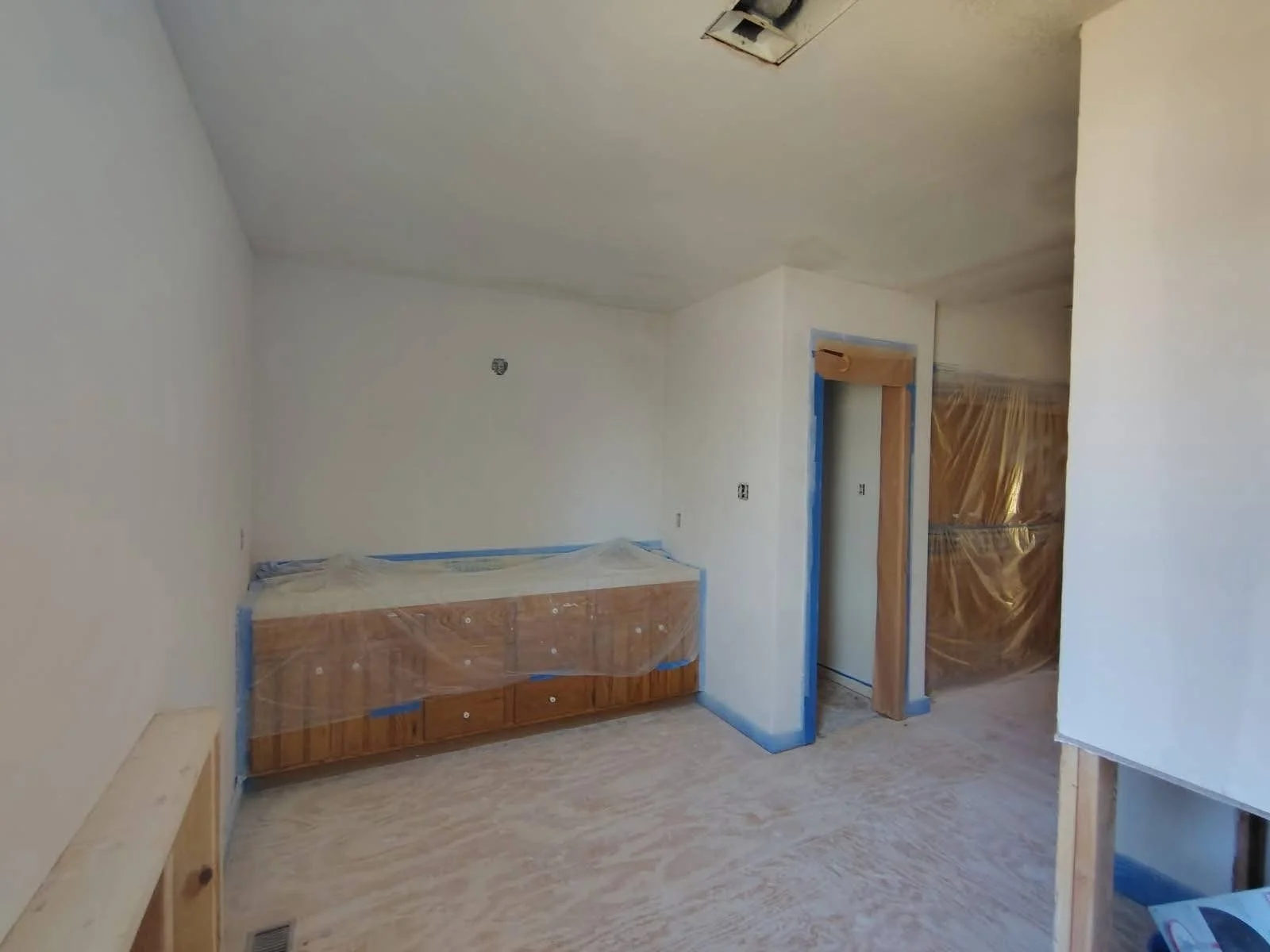 Room under construction with light-colored walls, a wooden cabinet covered with plastic, a doorway framed with blue tape, and plastic covering over another entrance. The flooring appears unfinished.