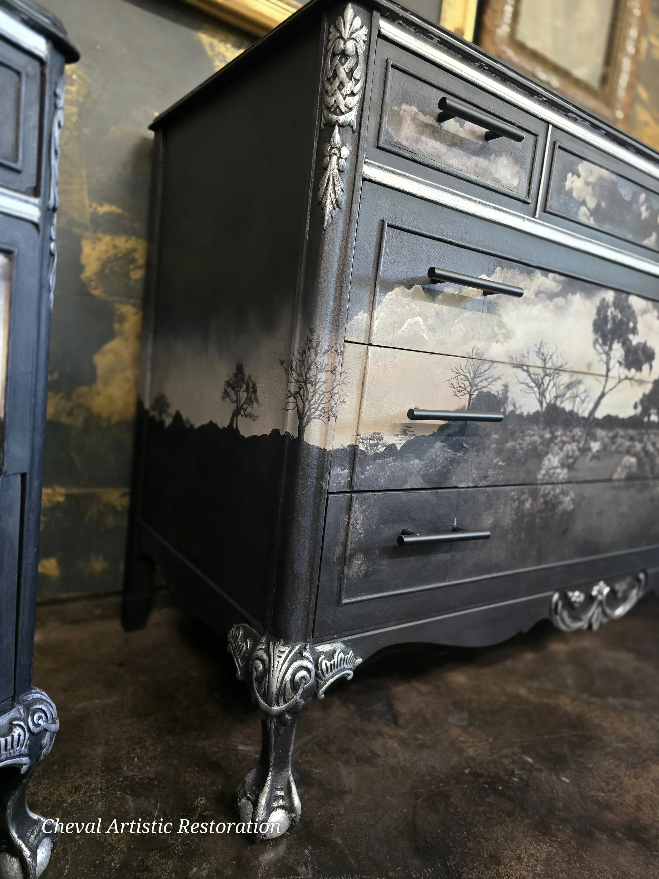 A black wooden dresser with ornate silver-colored carved details on the corners and feet, featuring a scenic landscape painting of trees and mountains on the front. The dresser has three drawers with black handles, and it is placed on a dark floor wi