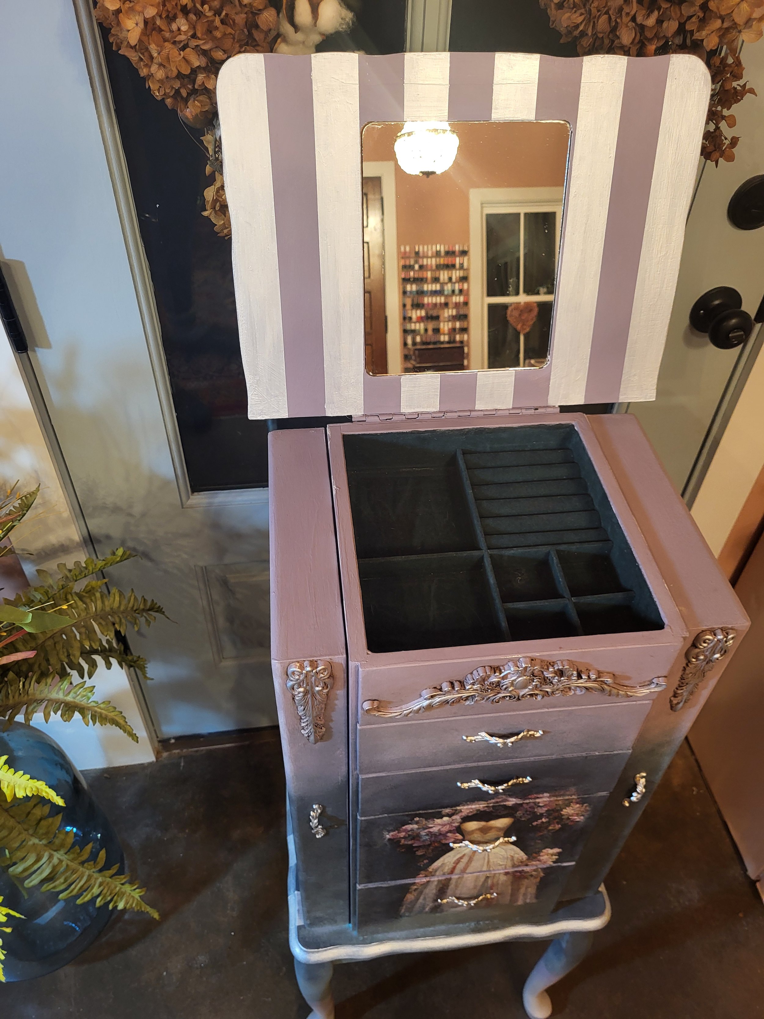 A vintage-style jewelry box with a mirror on the inside of the lid. The jewelry box is painted in pastel colors with ornate gold accents and vintage illustrations on the front. The interior has several compartments for organizing jewelry.