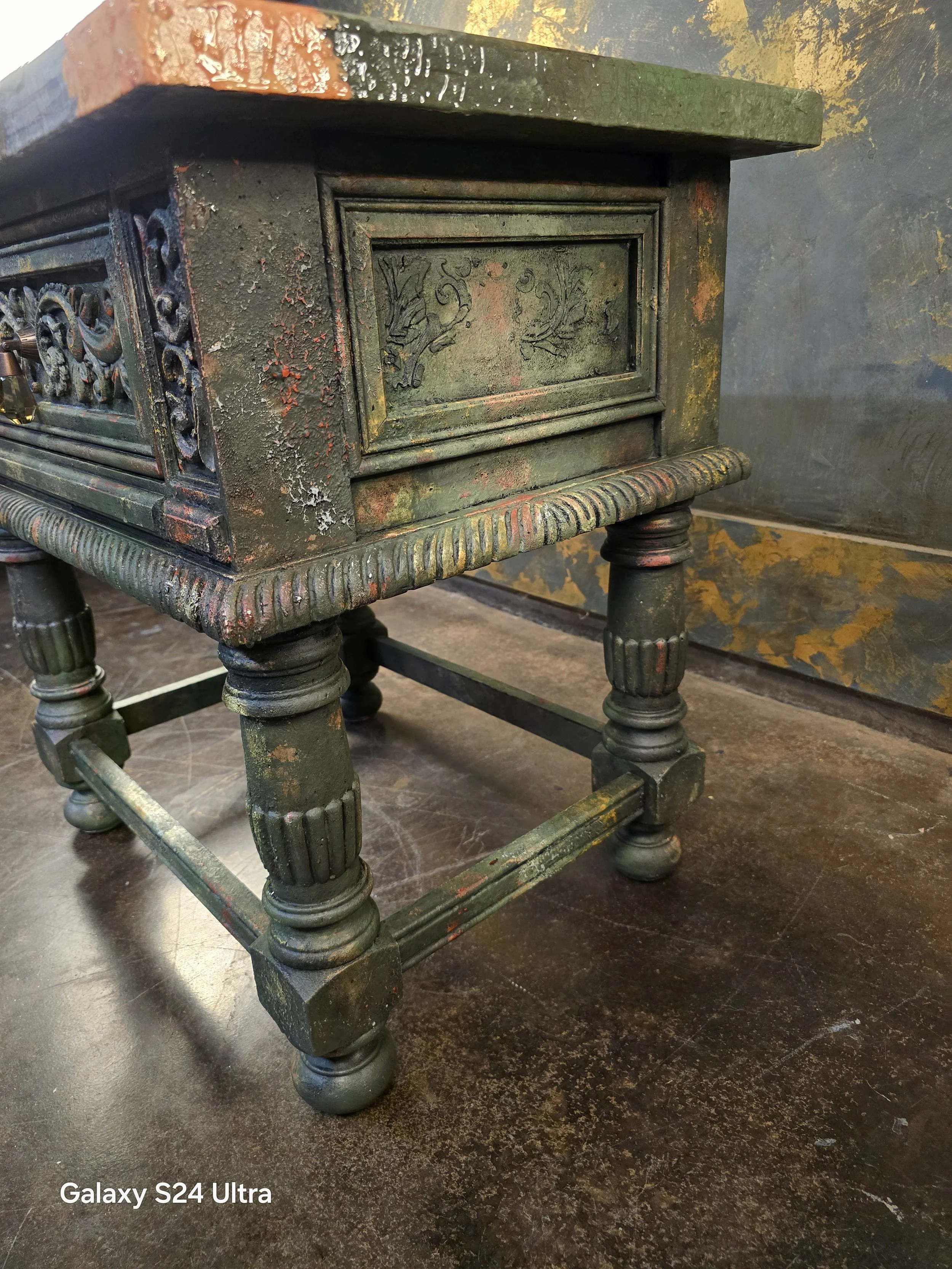 A vintage wooden side table with carved details, painted in dark green with a distressed finish, sits on a dark floor against an aged wall.