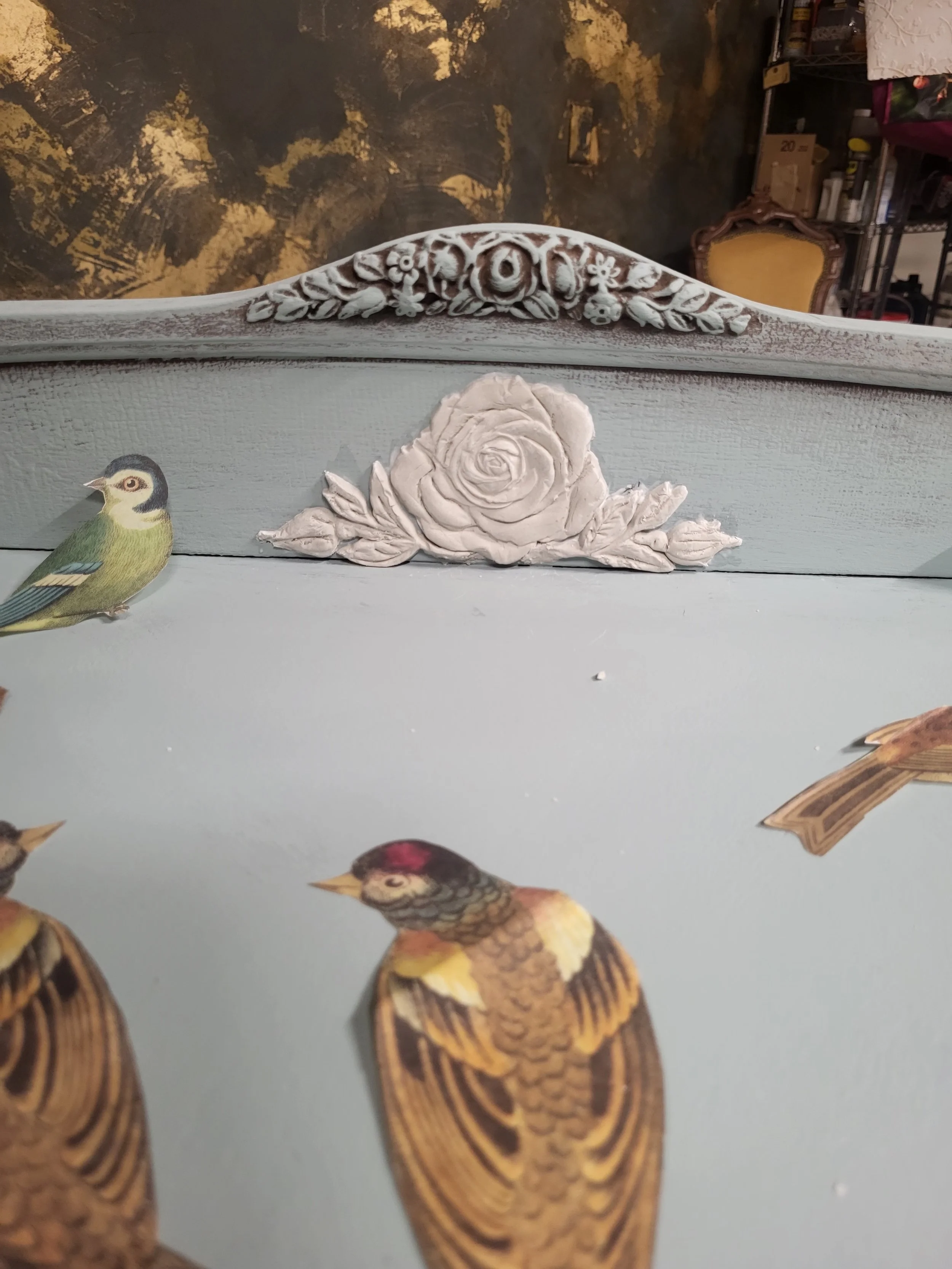 Close-up of a decorative piece with a painted bird design and a white carved rose with leaves on a pale blue background.