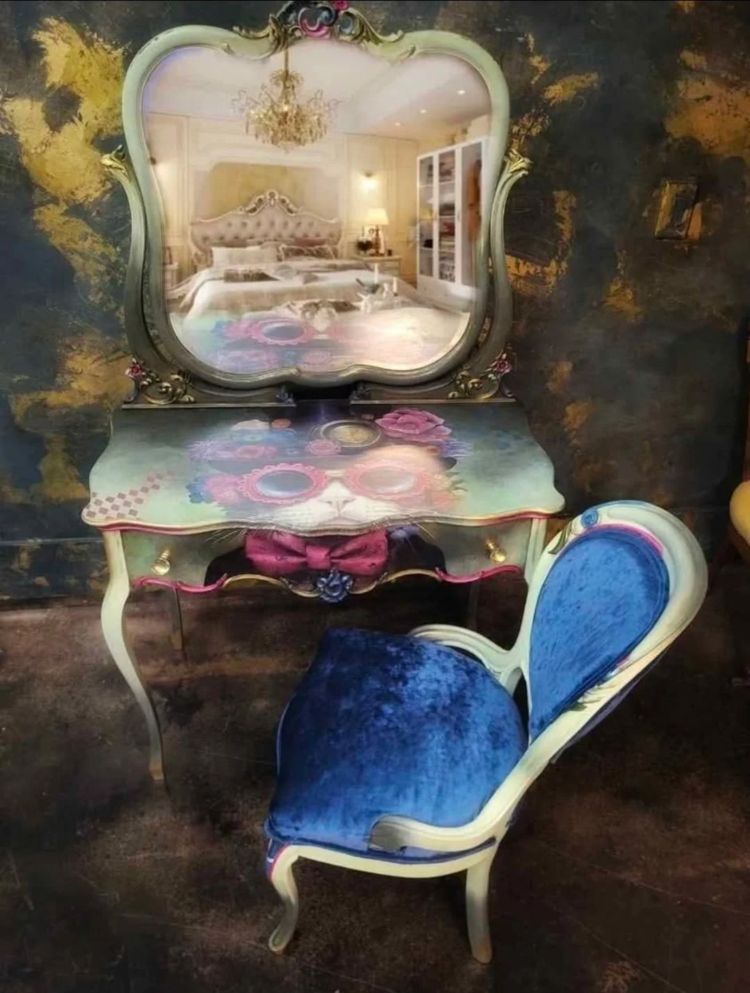 A vintage vanity table with a mirror, colorful floral design, and a blue velvet chair in front of it. The mirror reflects a bedroom with a bed, chandelier, and lamp.