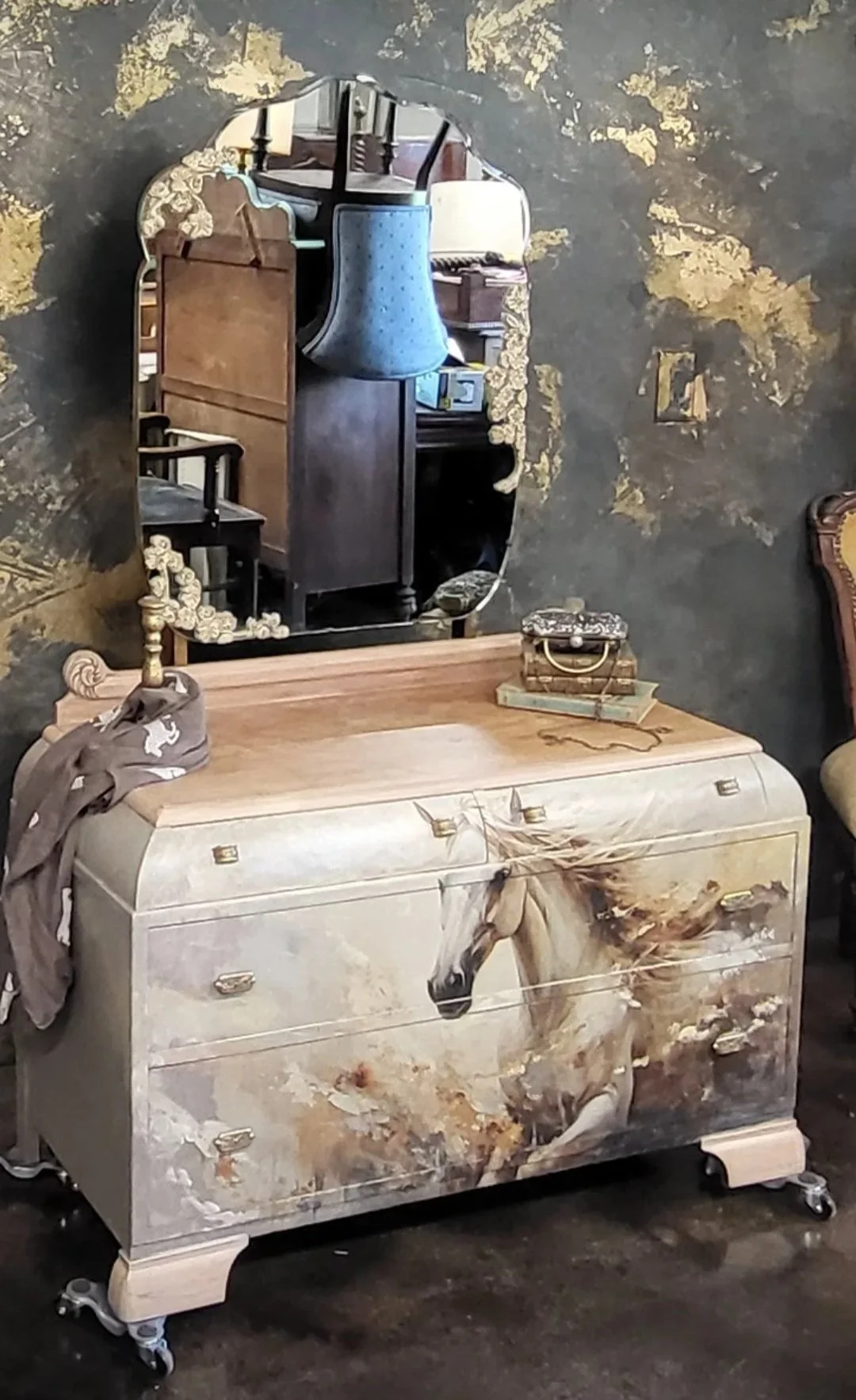 A vintage dresser with a painted horse design on the front, topped with a wooden surface, in a room with a dark textured wall and a mirror reflecting various furniture pieces.