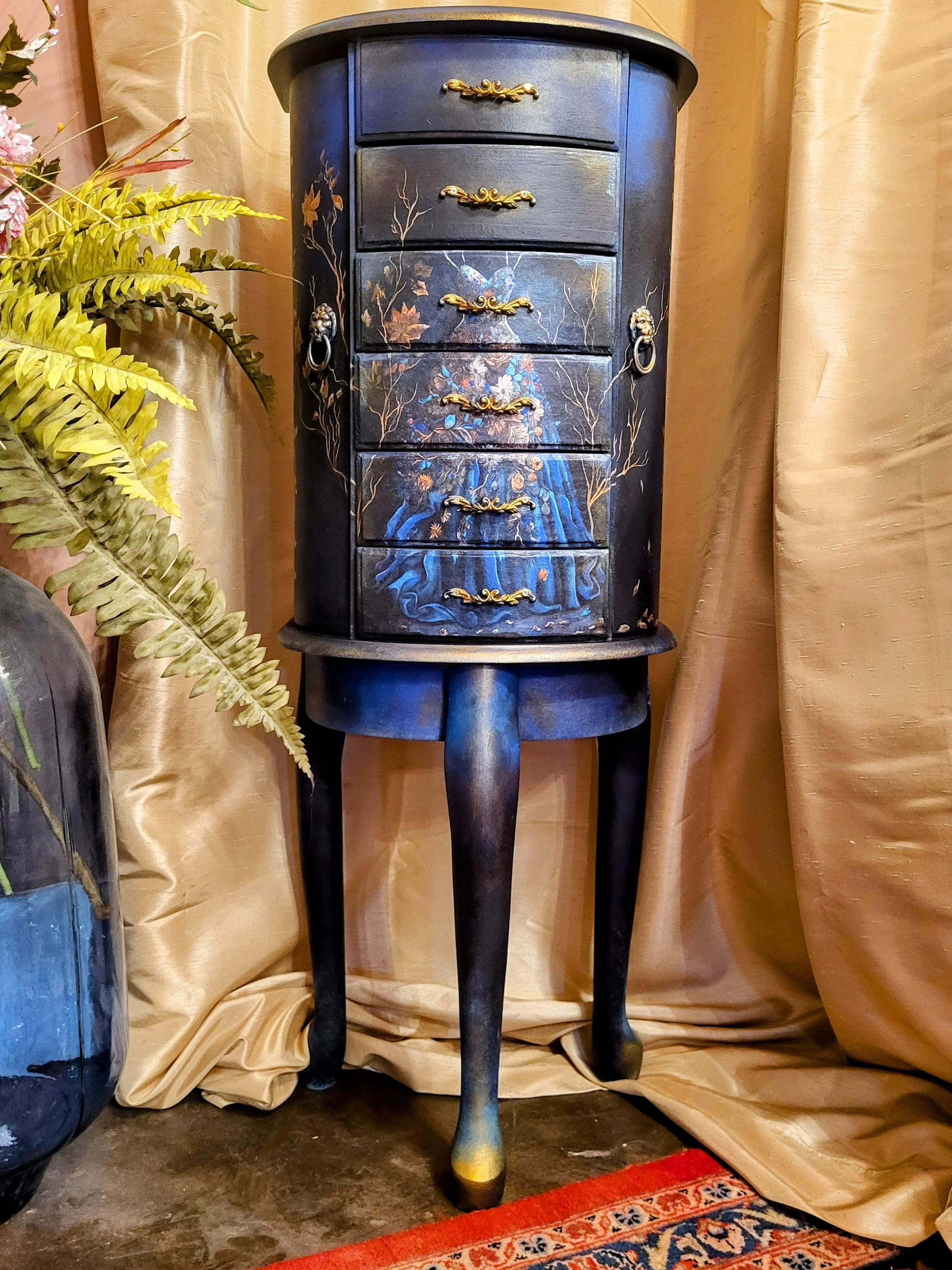 A black-painted wooden cabinet with floral and butterfly designs and gold handles, placed on a tall, black, curved-legged stand, with beige curtains and a potted plant nearby.