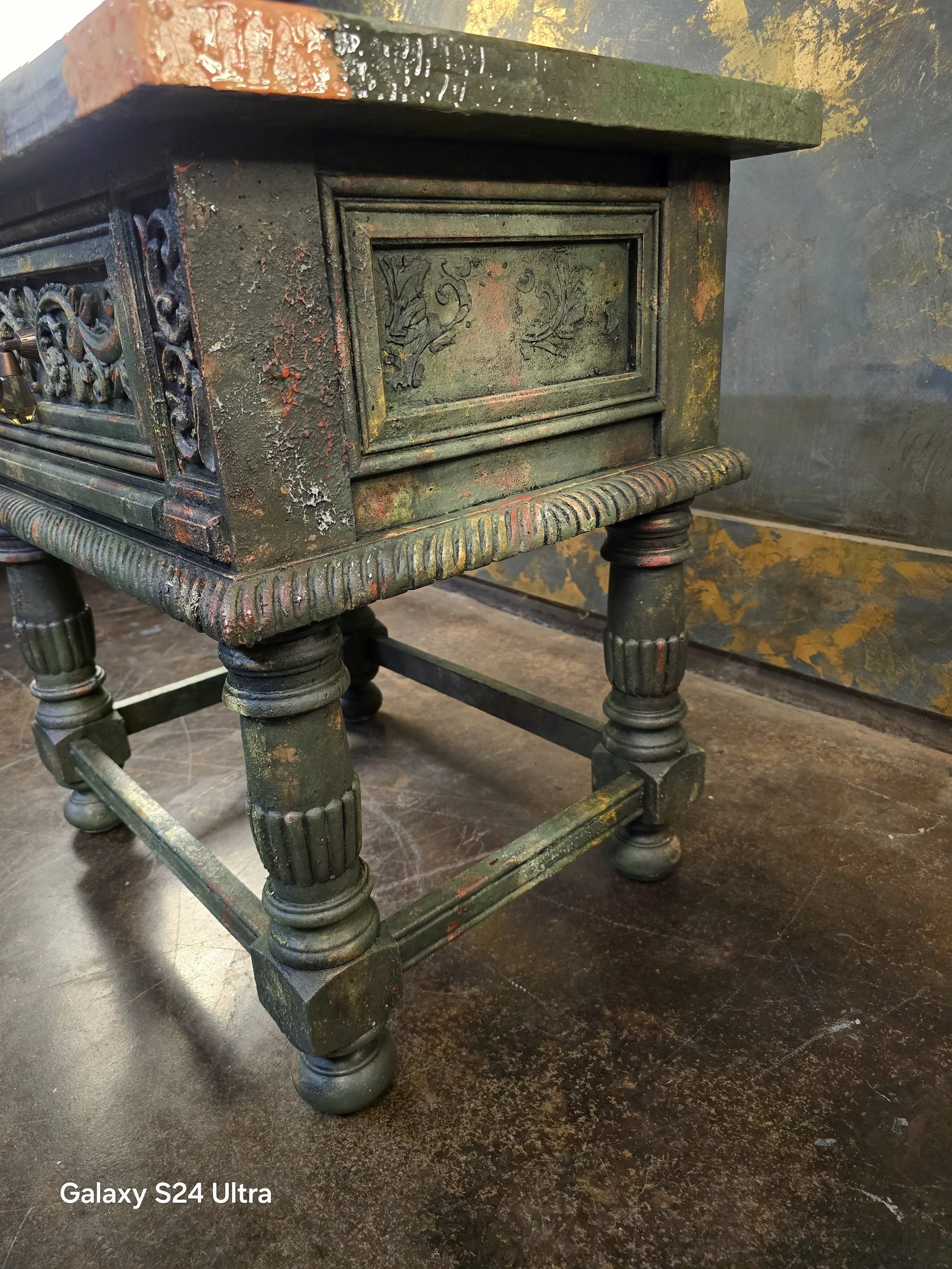 A close-up of an antique green-painted wooden table with ornate carvings, turned legs, and a distressed finish showing patches of exposed wood and worn paint.