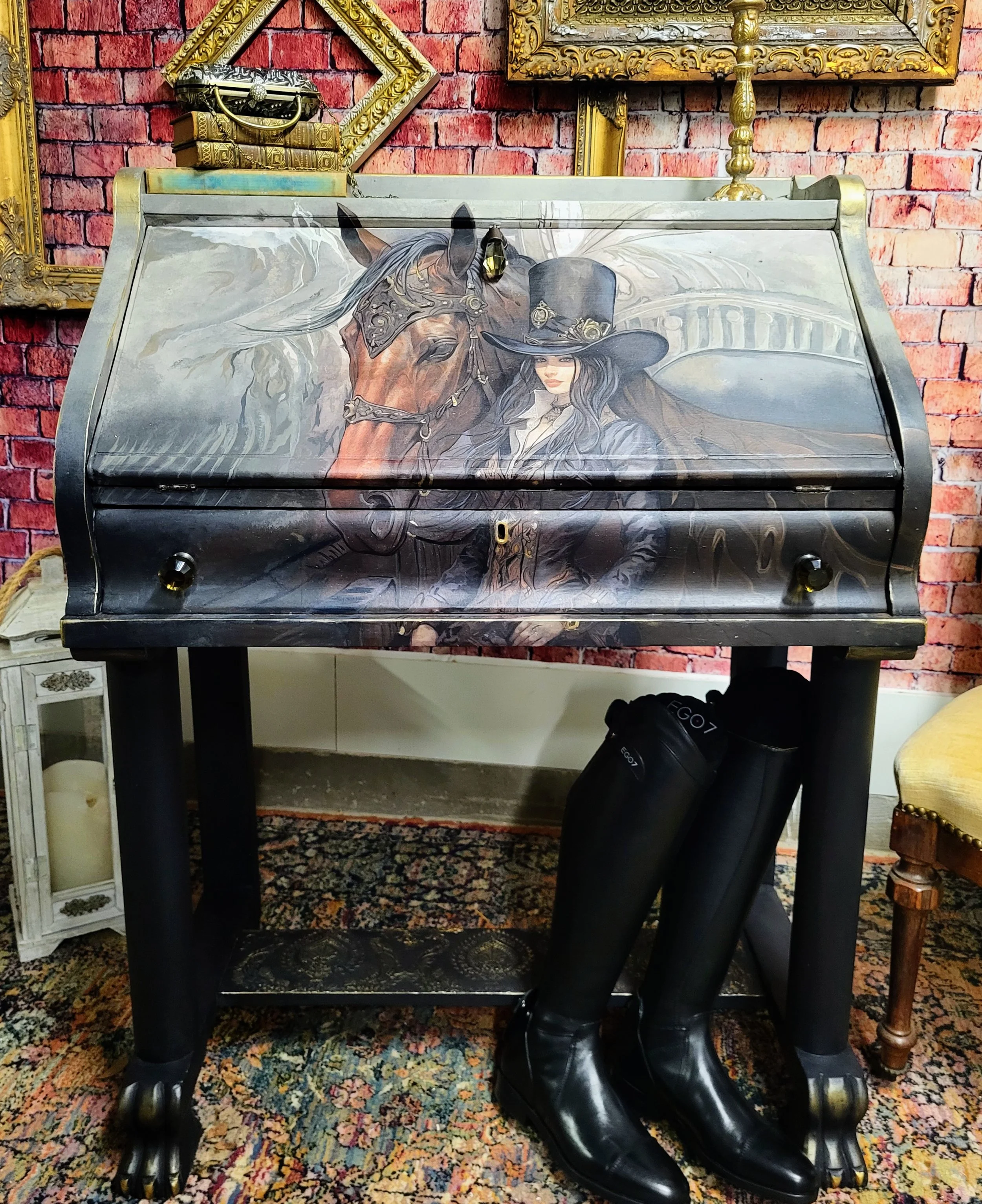 Decorative jewelry box with a painted image of a woman wearing a top hat and a horse, set against a brick wall. The jewelry box is on a black table, with tall black riding boots hanging on the side.