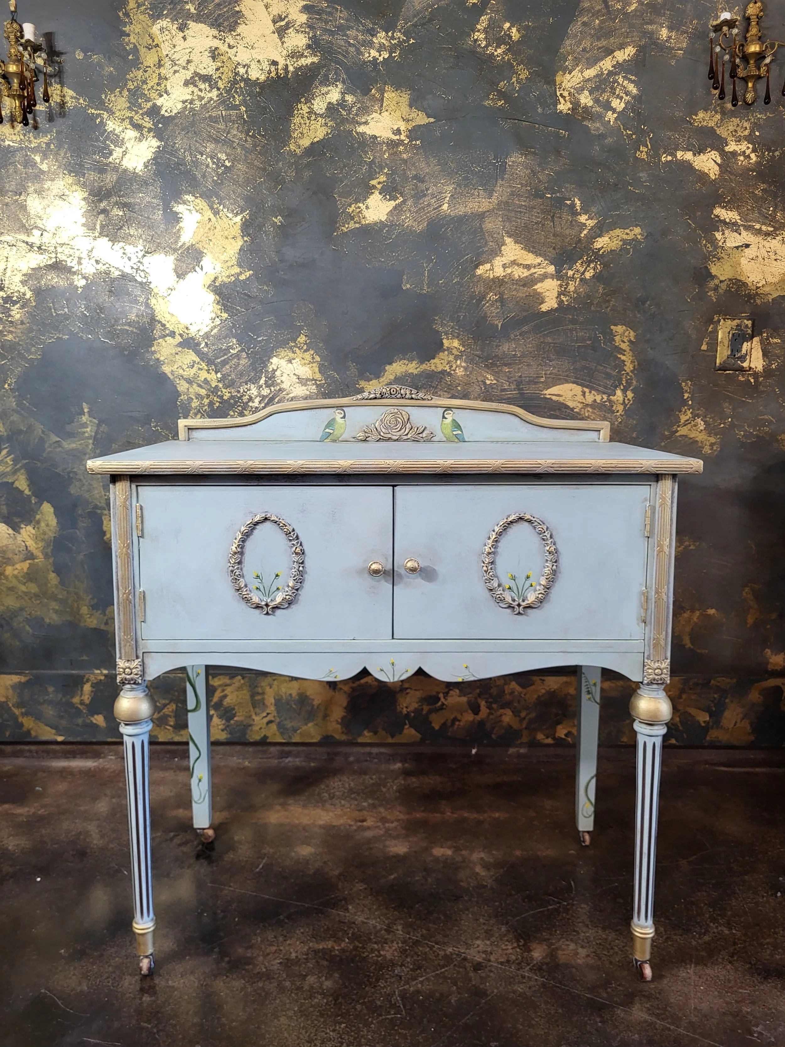 A vintage light blue wooden cabinet with painted details of birds and flowers, gold accents, and ornate handles, standing against a dark, gold-speckled wall.
