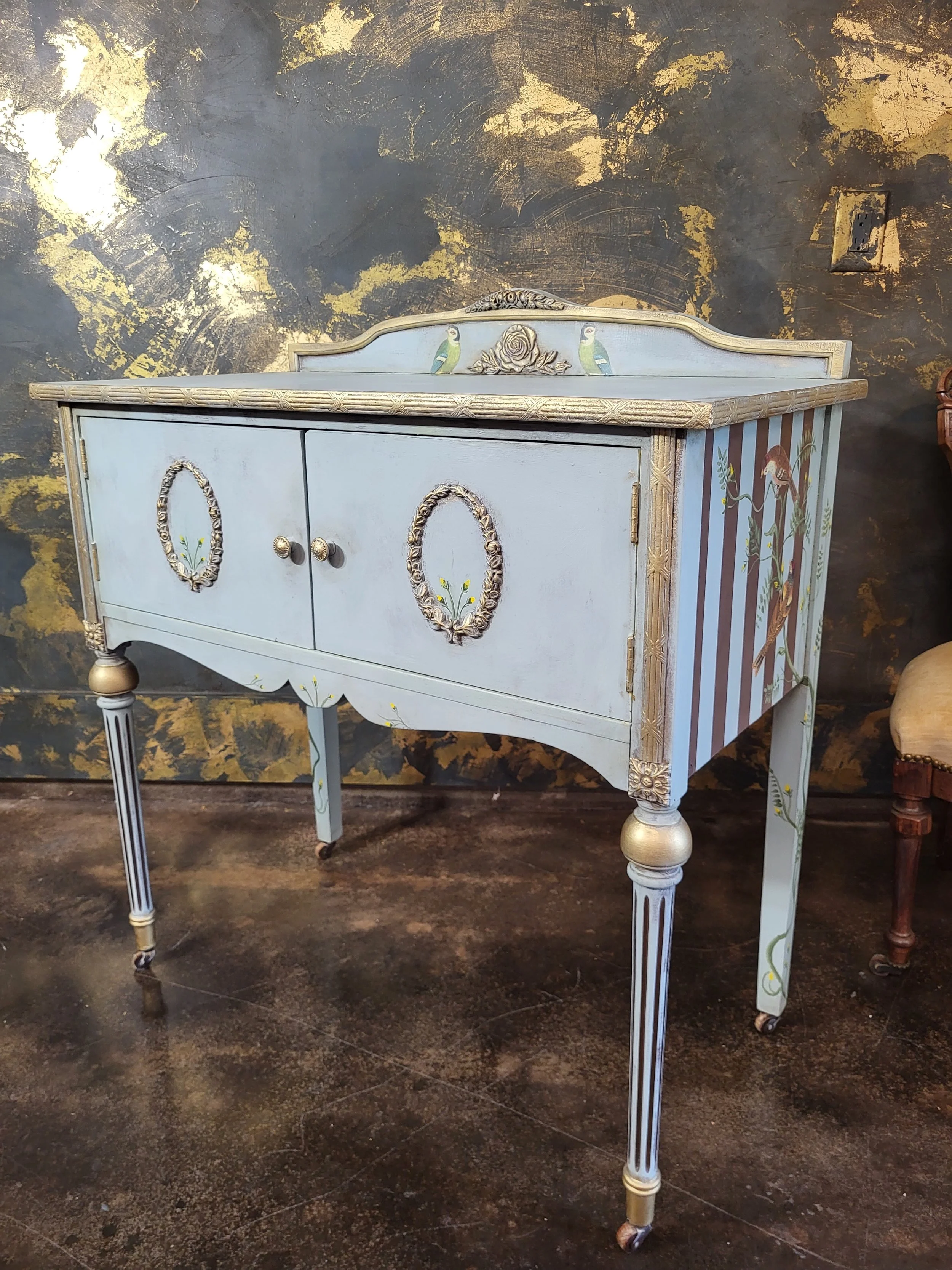 A vintage light blue wooden sideboard or cabinet with ornate gold and floral accents. It features two doors with decorative oval wreaths and is supported by four slender, carved legs.