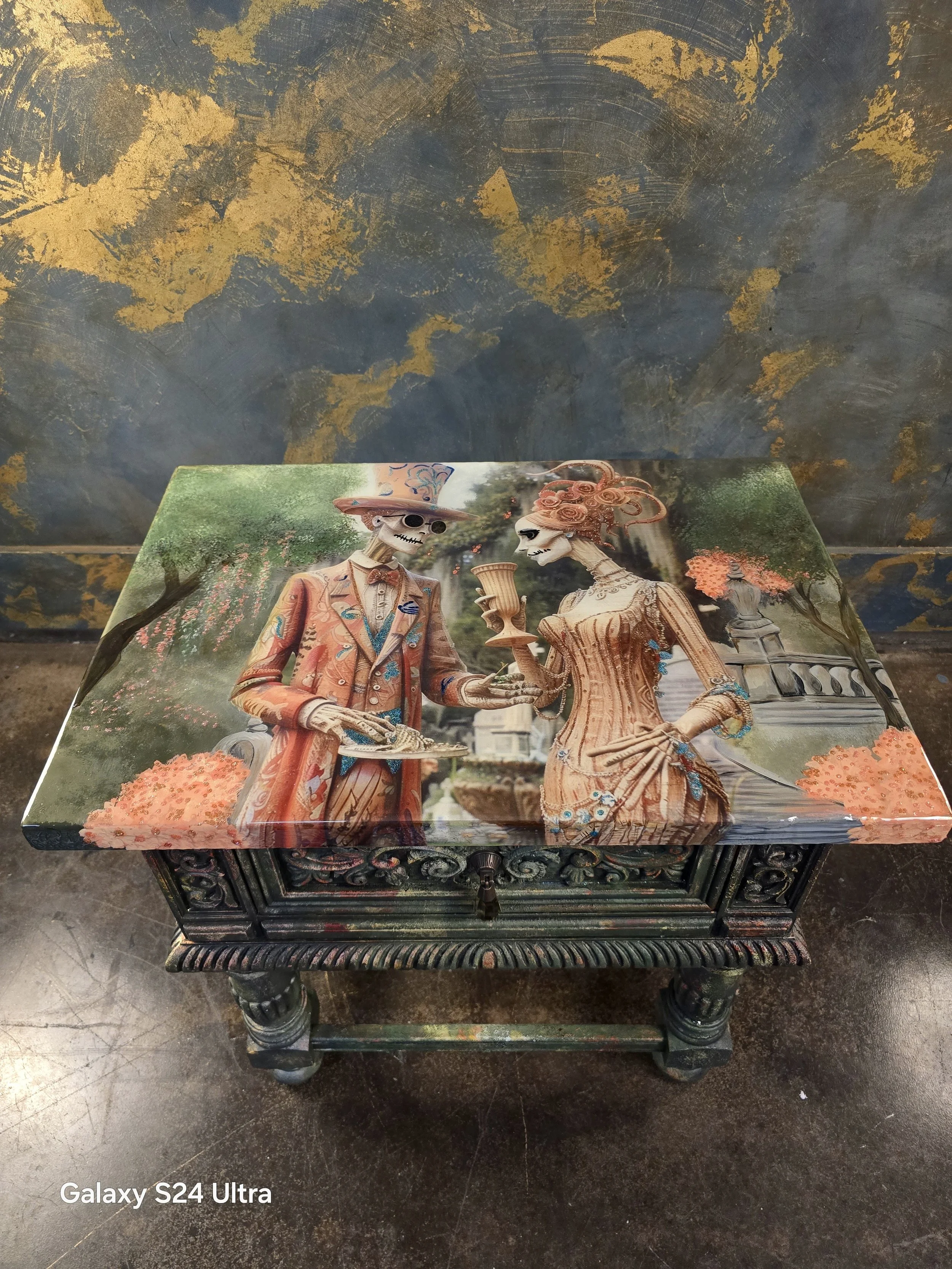 Decorative chest featuring skeleton man and woman in ornate, vintage clothing, depicted in a Day of the Dead style, set in a garden with trees, flowers, and a fountain.