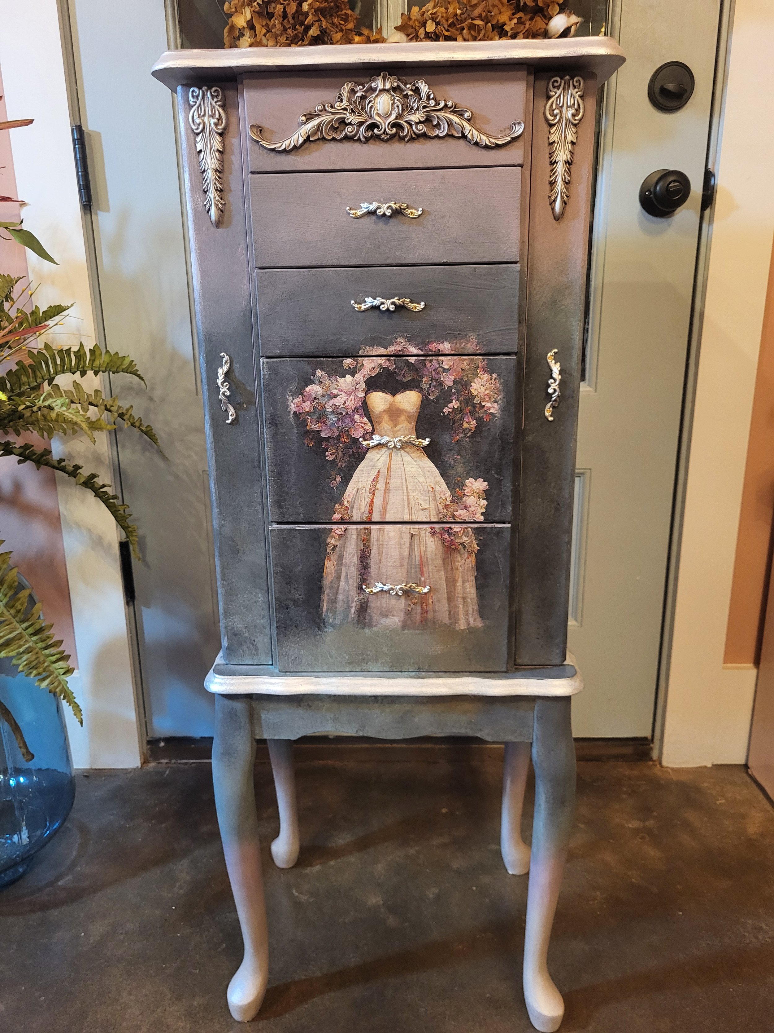 Decorative wooden cabinet with ornate silver handles and a floral dress painting on the front, standing on curved legs, placed indoors near a door and a potted plant.