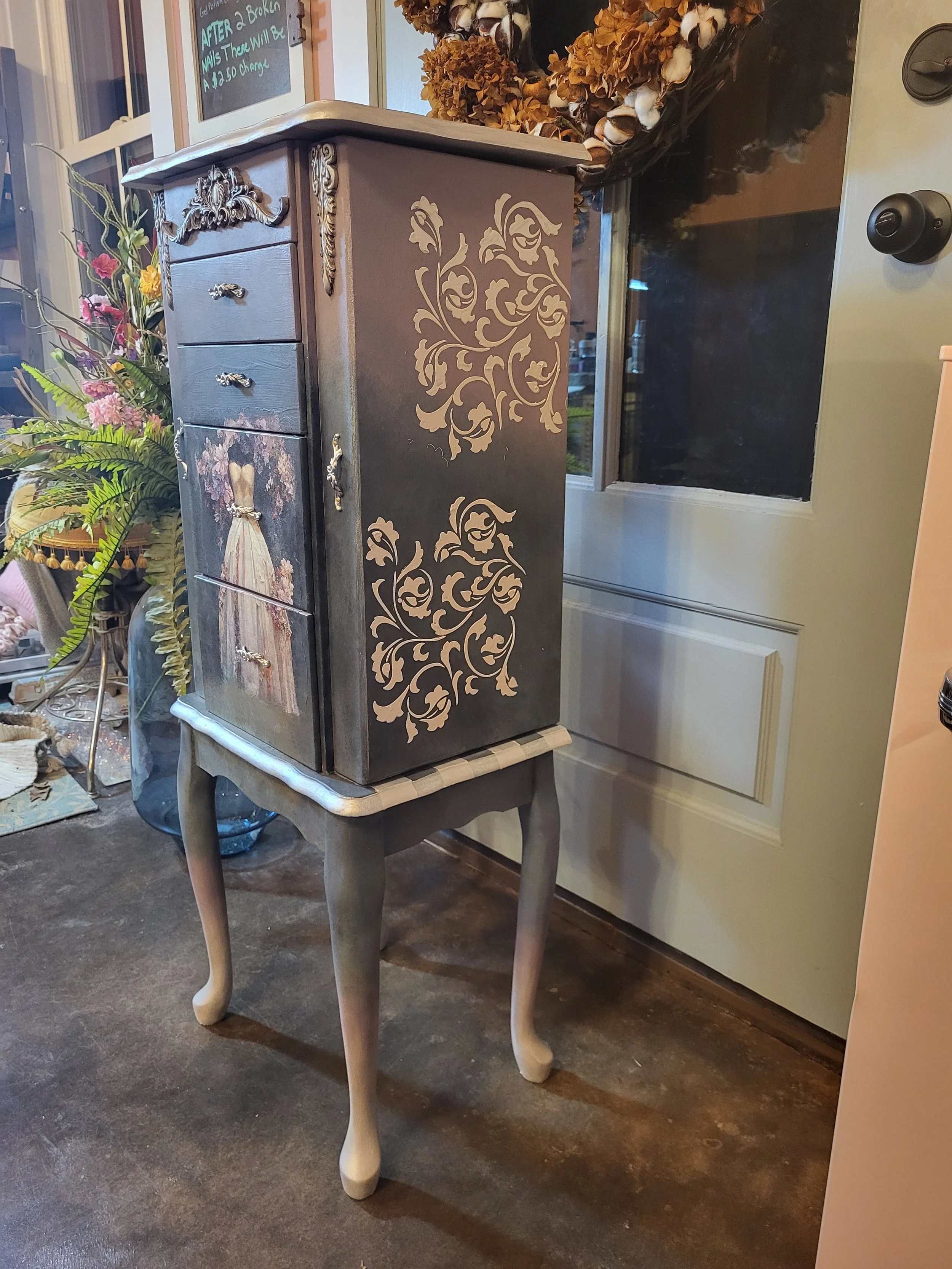A vintage-style wooden cabinet painted black with ornate gold floral designs on the side, placed on a small curved-legged wooden table, with a floral dress painting on the front panel, in a room decorated with flowers and a cotton wreath on the wall.
