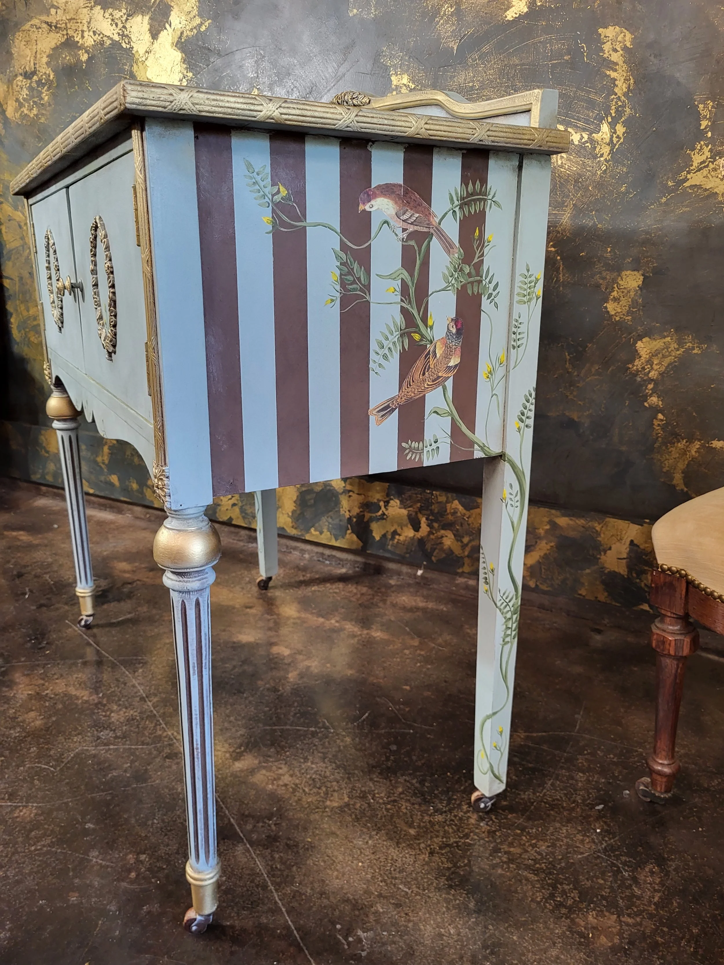Decorative antique side table with painted bird motif on the front panel, featuring two birds perched on branches, set against a background of vertical stripes in muted colors, with a carved edging on the top and slender, painted legs.