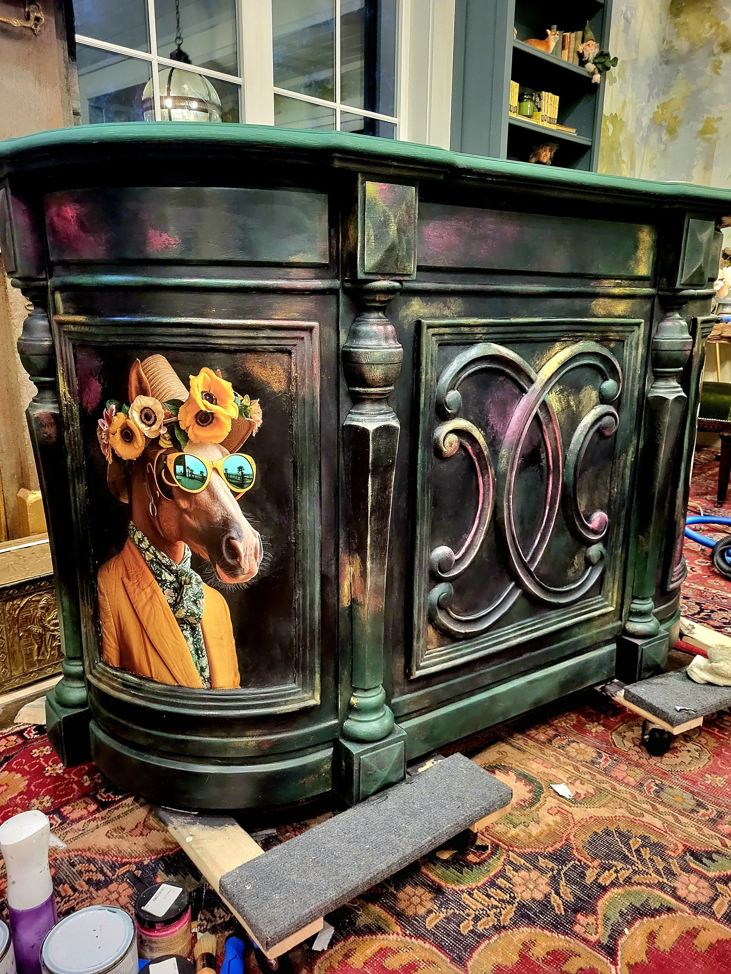 An ornate dark green cabinet with a painted fruit motif on the doors. There is a ledge with a dog sculpture, dressed in yellow and green clothing, with a sunflower hat and sunglasses, standing to the left of the cabinet.