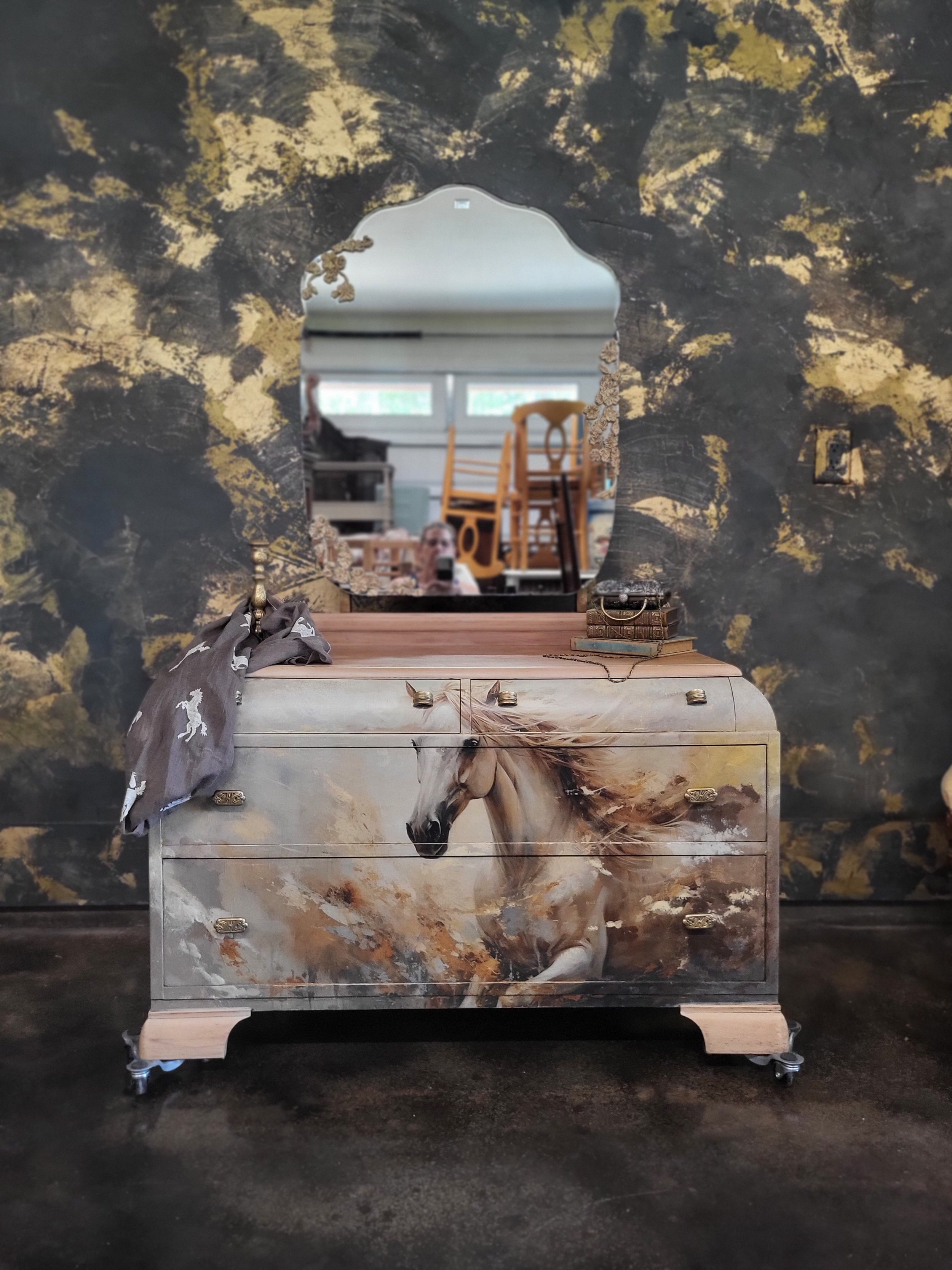 A vintage dresser with a painted horse design on the front, topped with a mirror, a gray scarf, a small stack of books, and a few other small items. The background features a dark textured wall with gold accents.