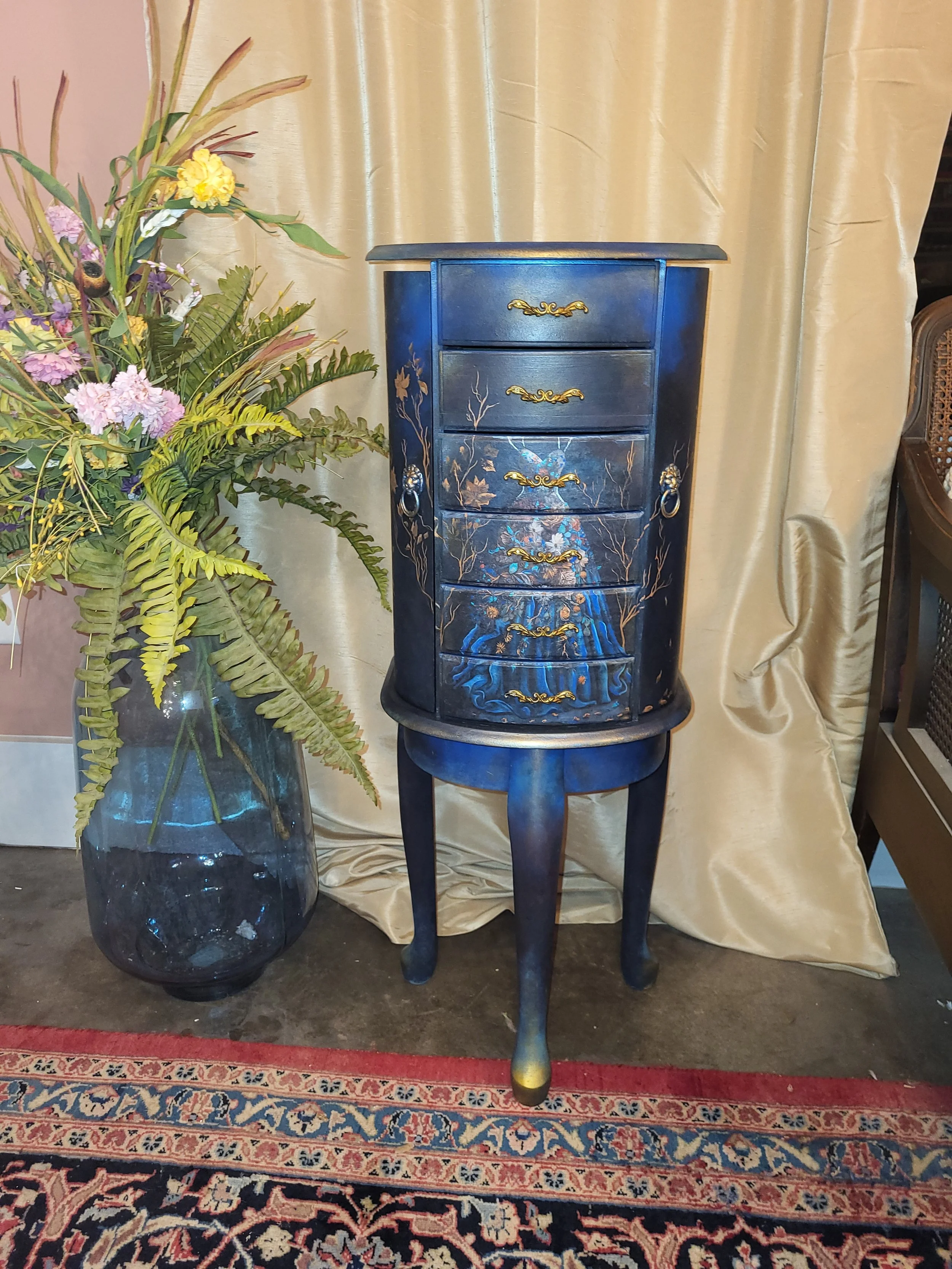 A tall, round, dark blue cabinet with painted crane and tree designs, gold-handled drawers, and standing on curved legs next to a large glass vase filled with pink, purple, and yellow flowers and lush greenery. The background features gold curtains a