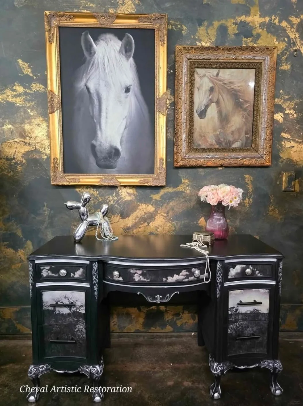 Interior scene with two framed horse portraits on a gold-leafed wall, a black and white ornate desk, a pink vase with flowers, a balloon sculpture of a dog, and jewelry.