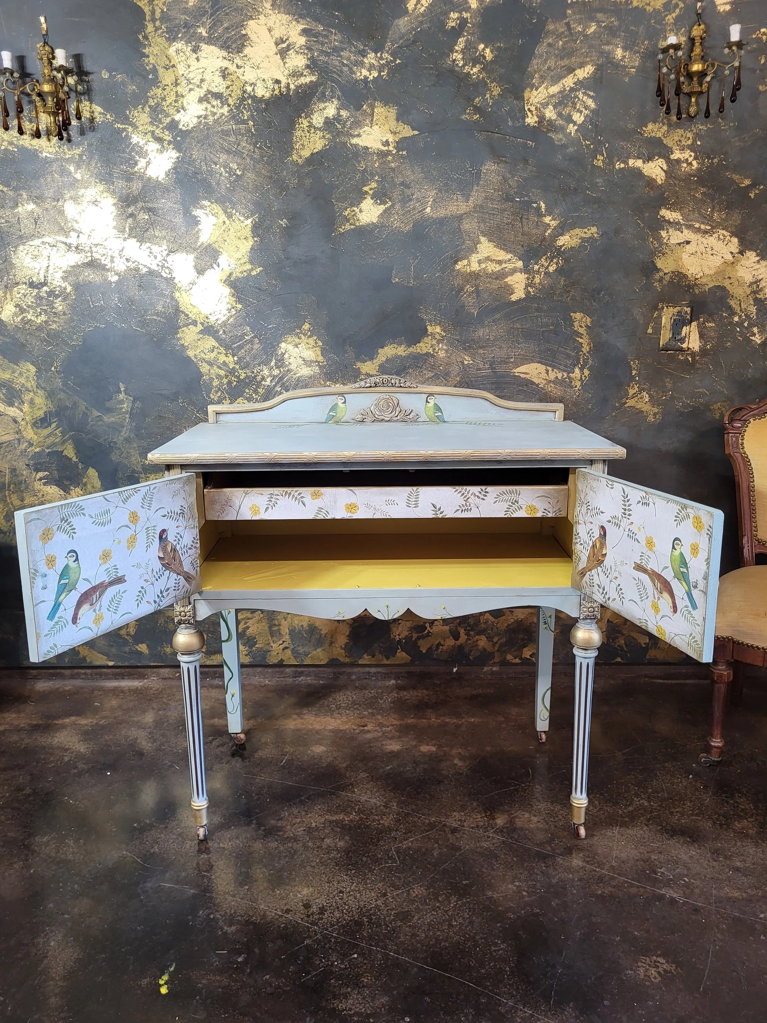 A vintage, painted wooden side table with decorative columns on the legs. The table has a light blue and white color scheme with bird and floral motifs, and features two side doors with similar designs. The table is set against a dark textured wall w