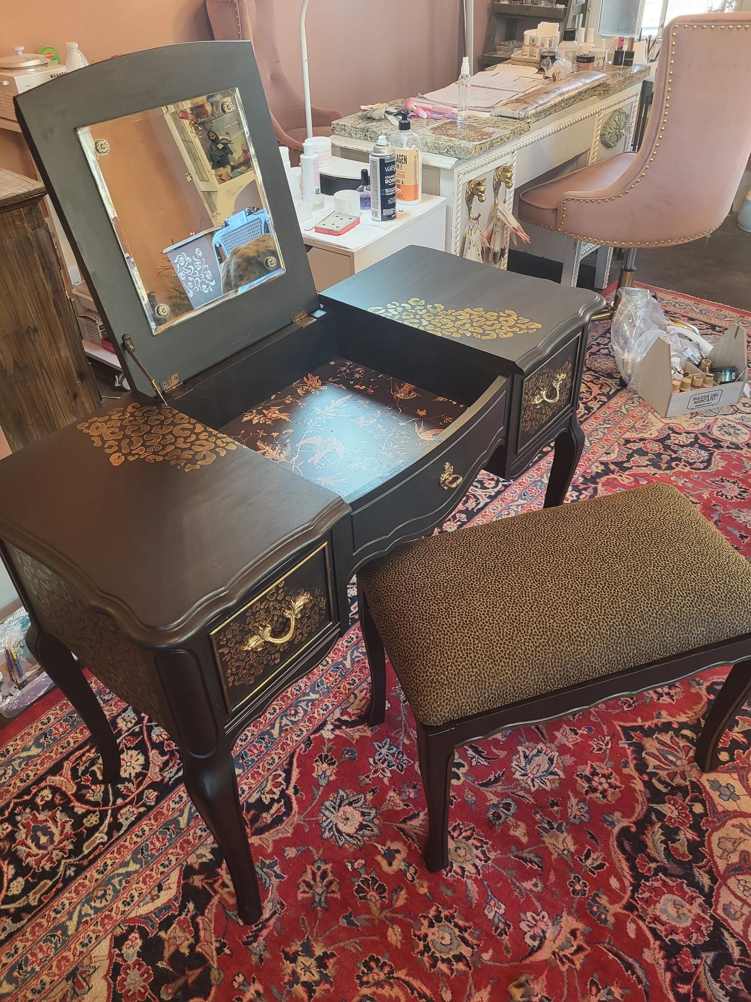 A black vintage jewelry box with gold accents and a mirror, placed on a richly patterned red and blue oriental rug. The box is open, showing compartments with gold leaf designs on the inner surfaces. A small green upholstered stool with black legs is