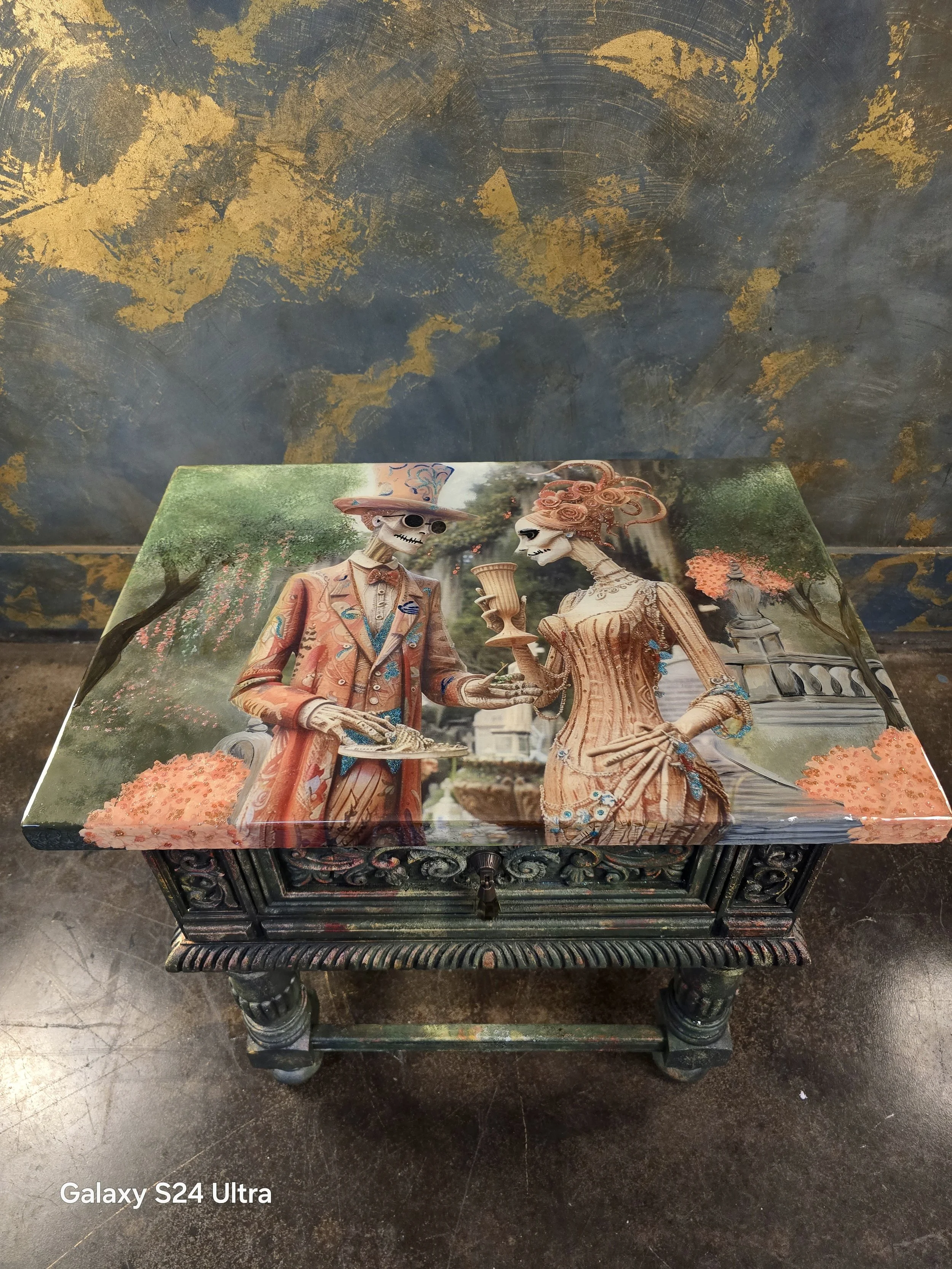 Decorative wooden box featuring painted skeleton figures in vintage clothing, set outdoors with greenery and flowers in the background.