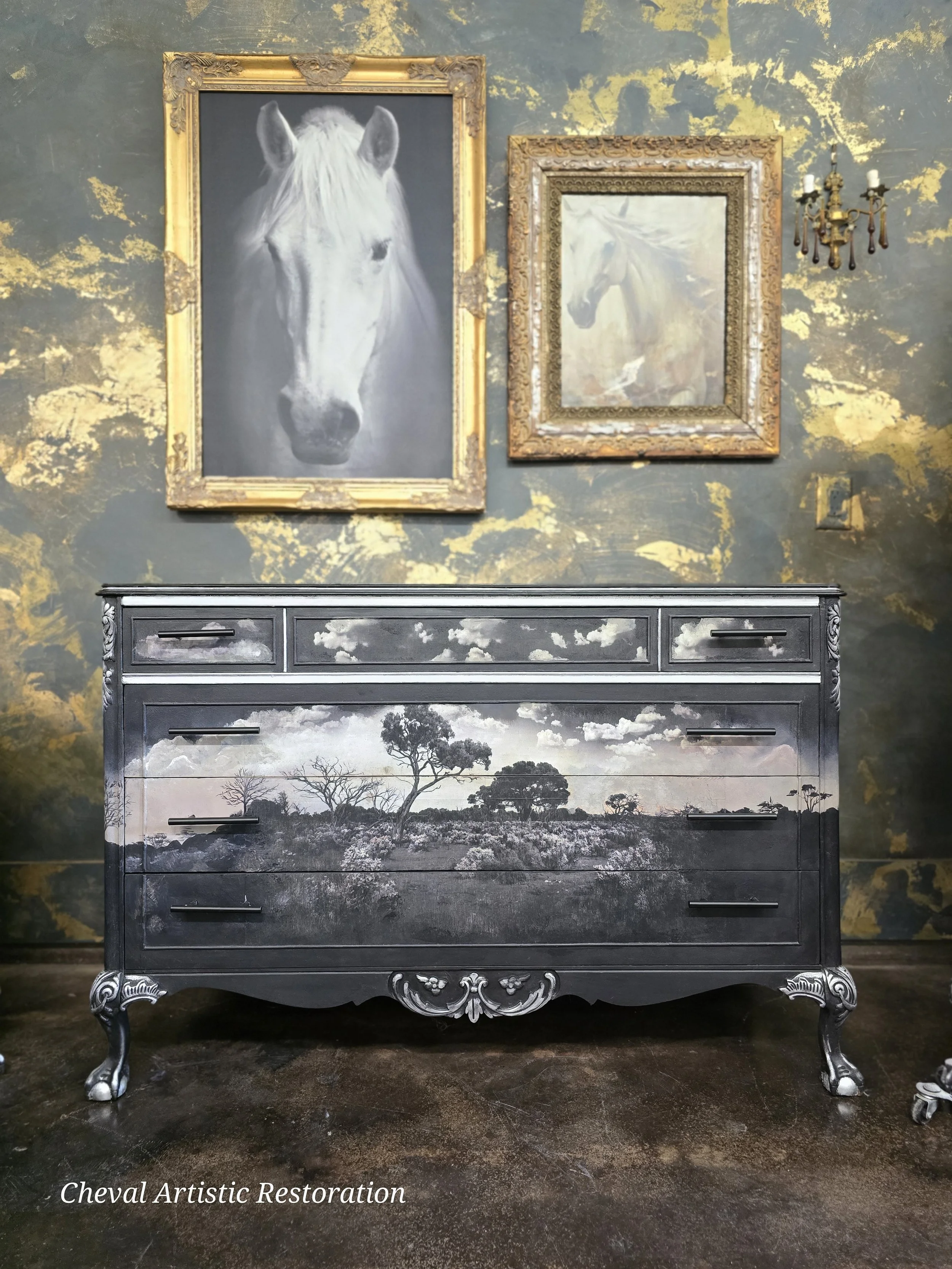 A black dresser with a landscape scene of trees and sky painted on the front, set against a wall with gold and dark gray textured paint. Above the dresser are two framed pictures of horses, one larger and painted in grayscale and the other smaller wi
