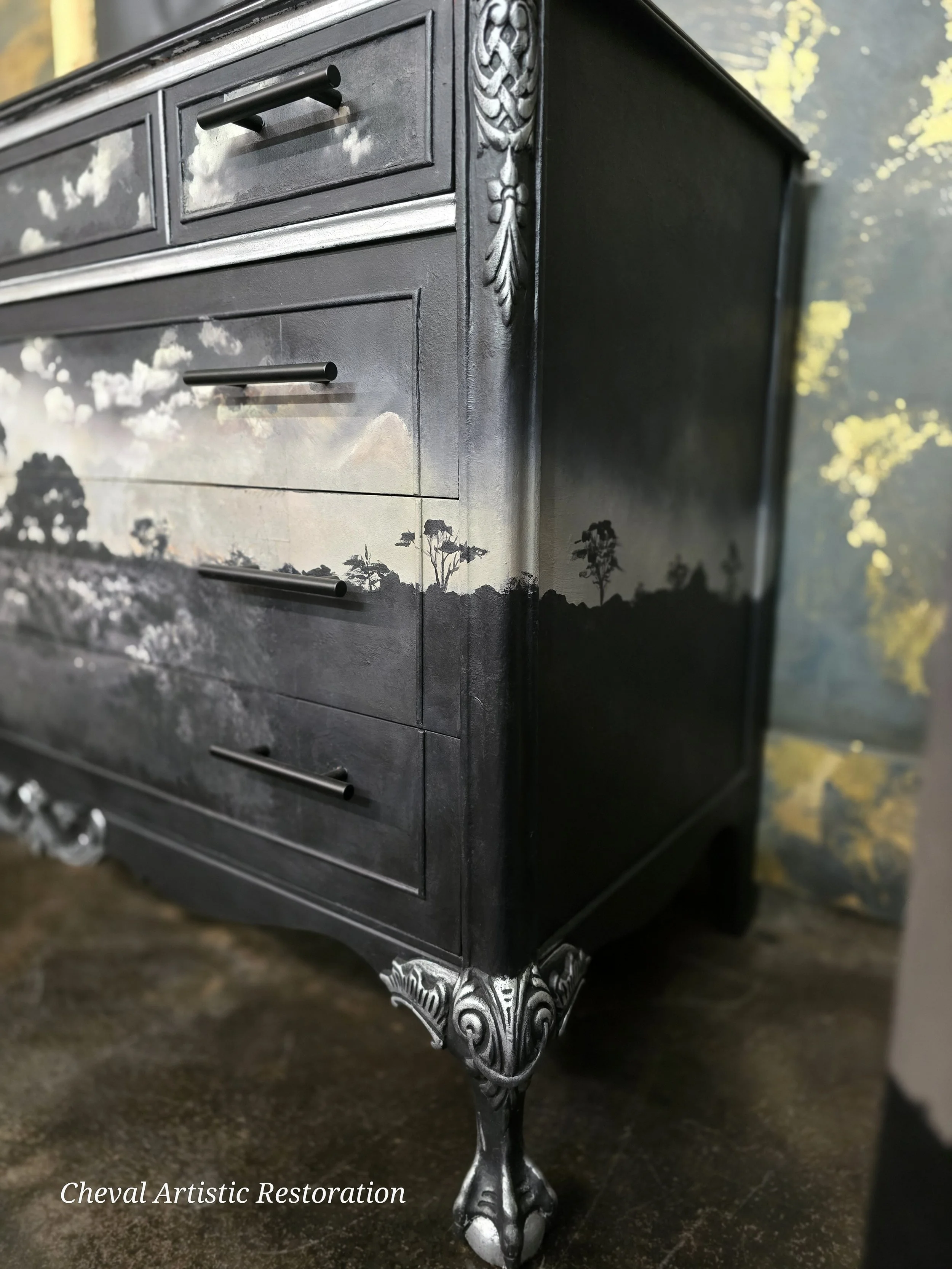 A black wooden dresser with floral and landscape painted designs, ornate silver-colored carved legs, and drawers with black handles. The background features a wall with a floral mural, and the dresser is placed on a dark floor.