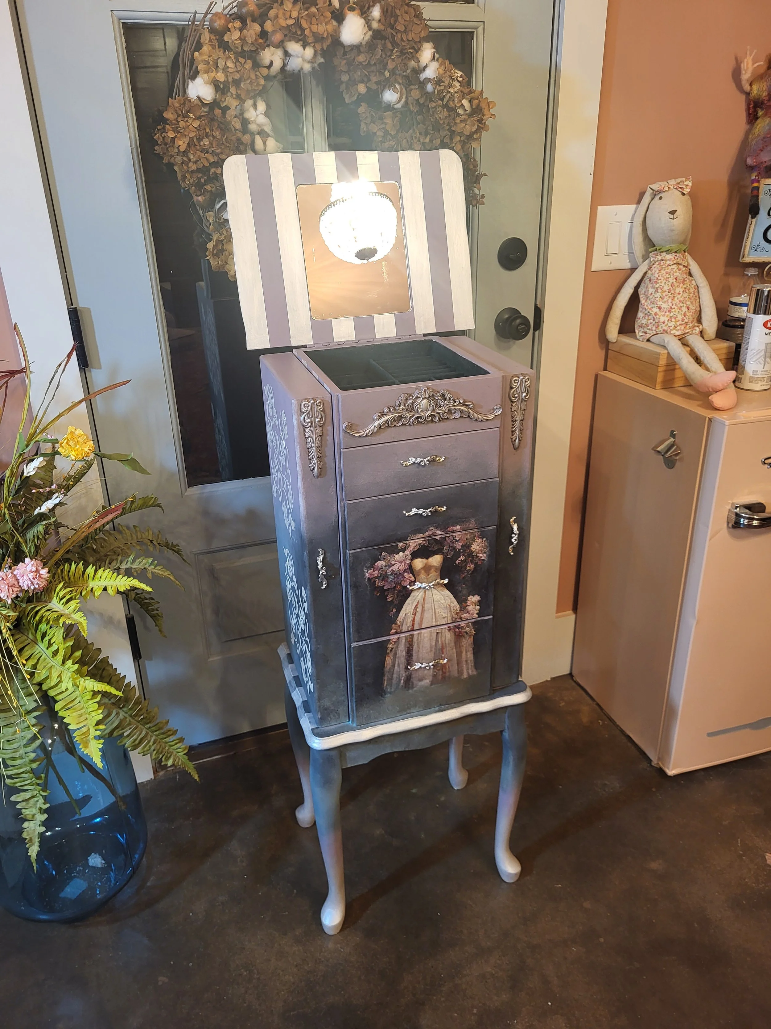 Decorative jewelry box with a hand-painted dress of a girl with flowers on the drawers, on a vintage-style legged stand, illuminated by a light, with a wreath hanging on a door behind.