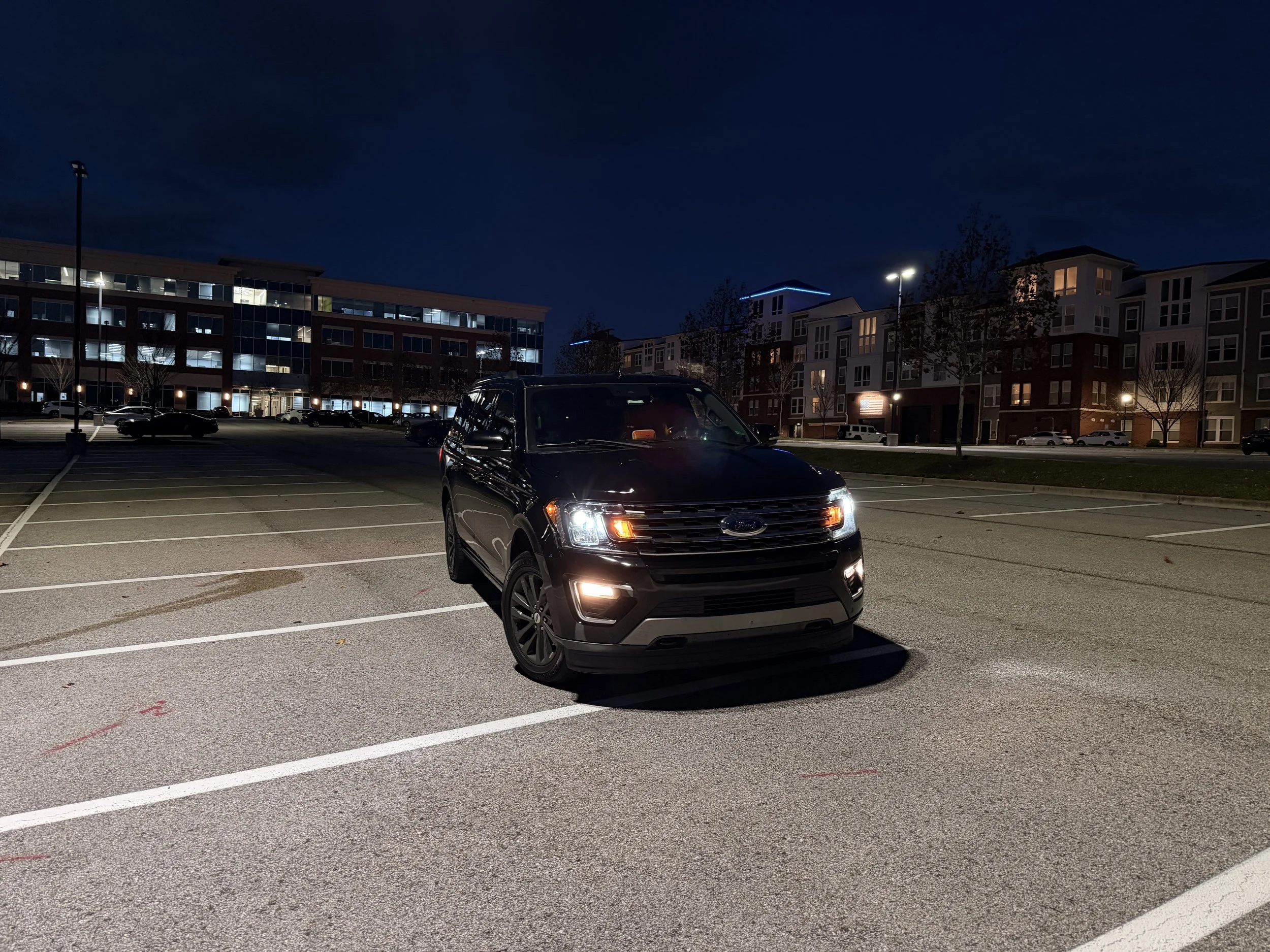 A black SUV parked in an outdoor parking lot at night, with buildings and streetlights in the background.