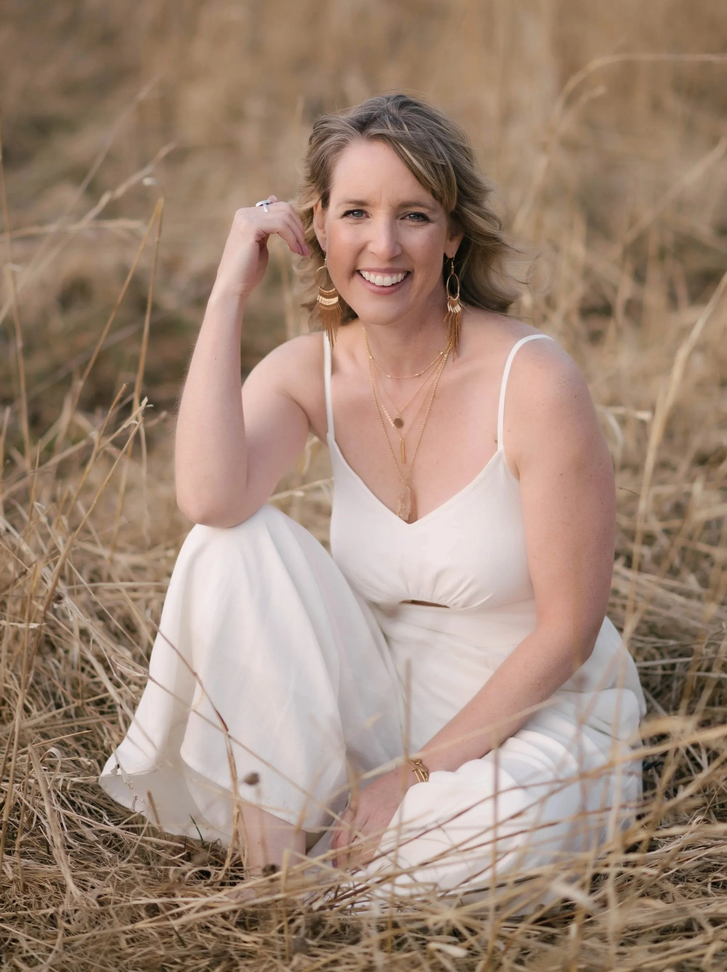 A woman sitting in tall, dry grass, smiling at the camera, wearing a white dress and gold jewelry.
