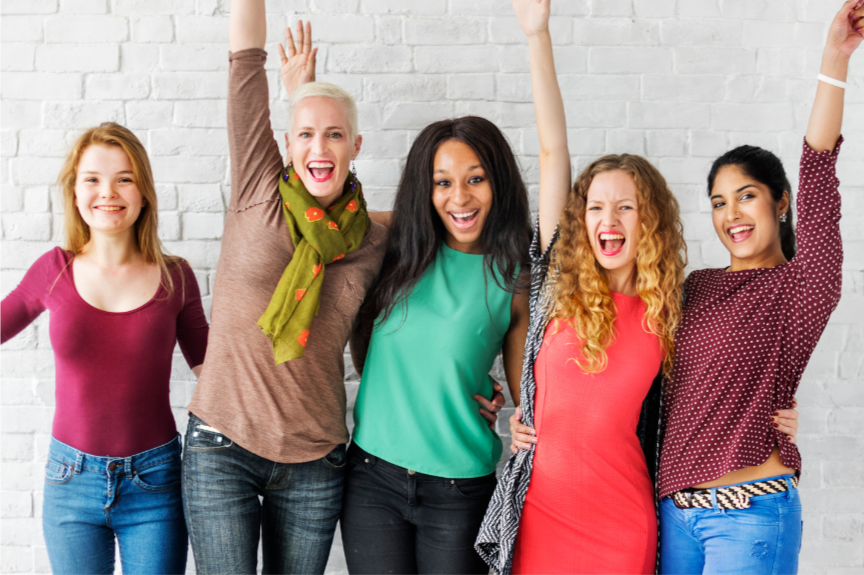 Five women standing together in front of a white brick wall, smiling, with two raising their hands in celebration.