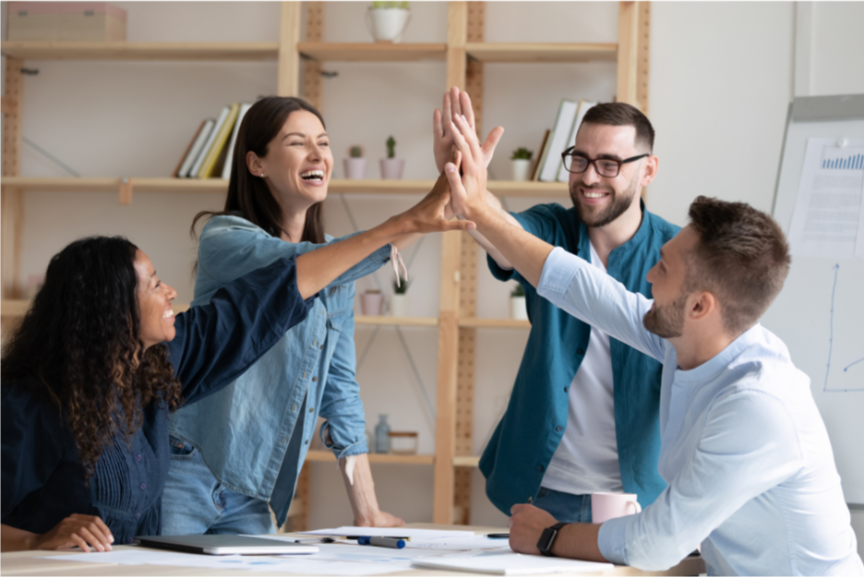 Four young professionals celebrating in a modern office with bookshelves and a chart in the background.
