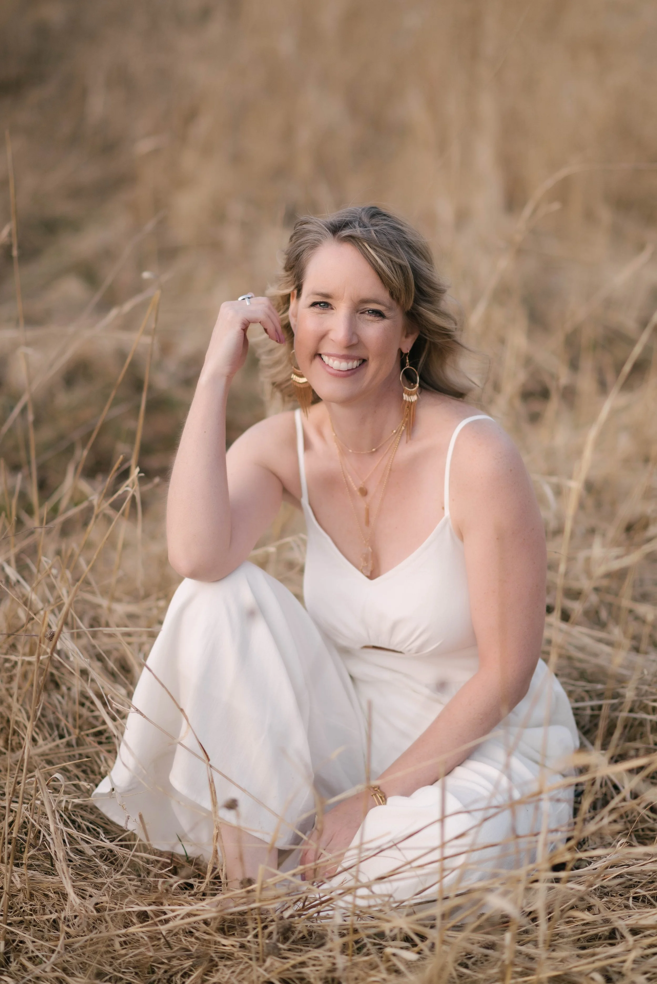 A woman with shoulder-length wavy hair, smiling and sitting on dry grass, dressed in a white sleeveless dress, accessorized with gold jewelry including earrings, layered necklaces, and a bracelet, in an outdoor setting with a blurred background of tall, dry grass.