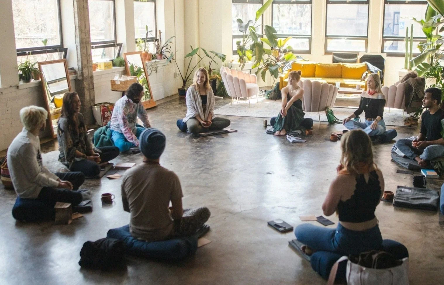 Group of people sitting in a circle on the floor in a bright, airy room with large windows and indoor plants, participating in a meditation or discussion session.